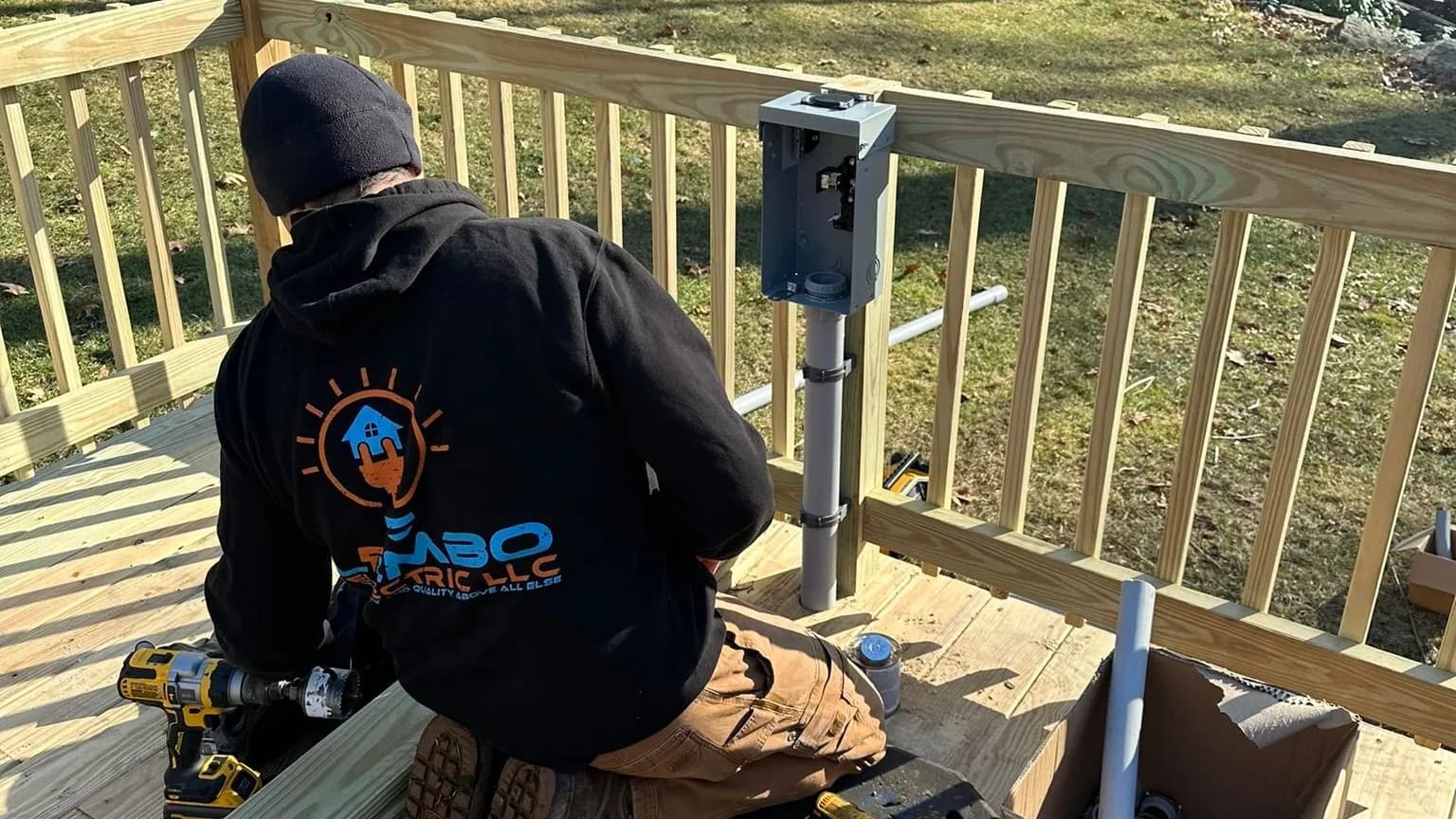 A technician in a black beanie and a hoodie with the Lembo logo, seen from behind, kneeling on a new deck beside a gray disconnect box mounted on a railing post with conduit, with tools and boxes around him.