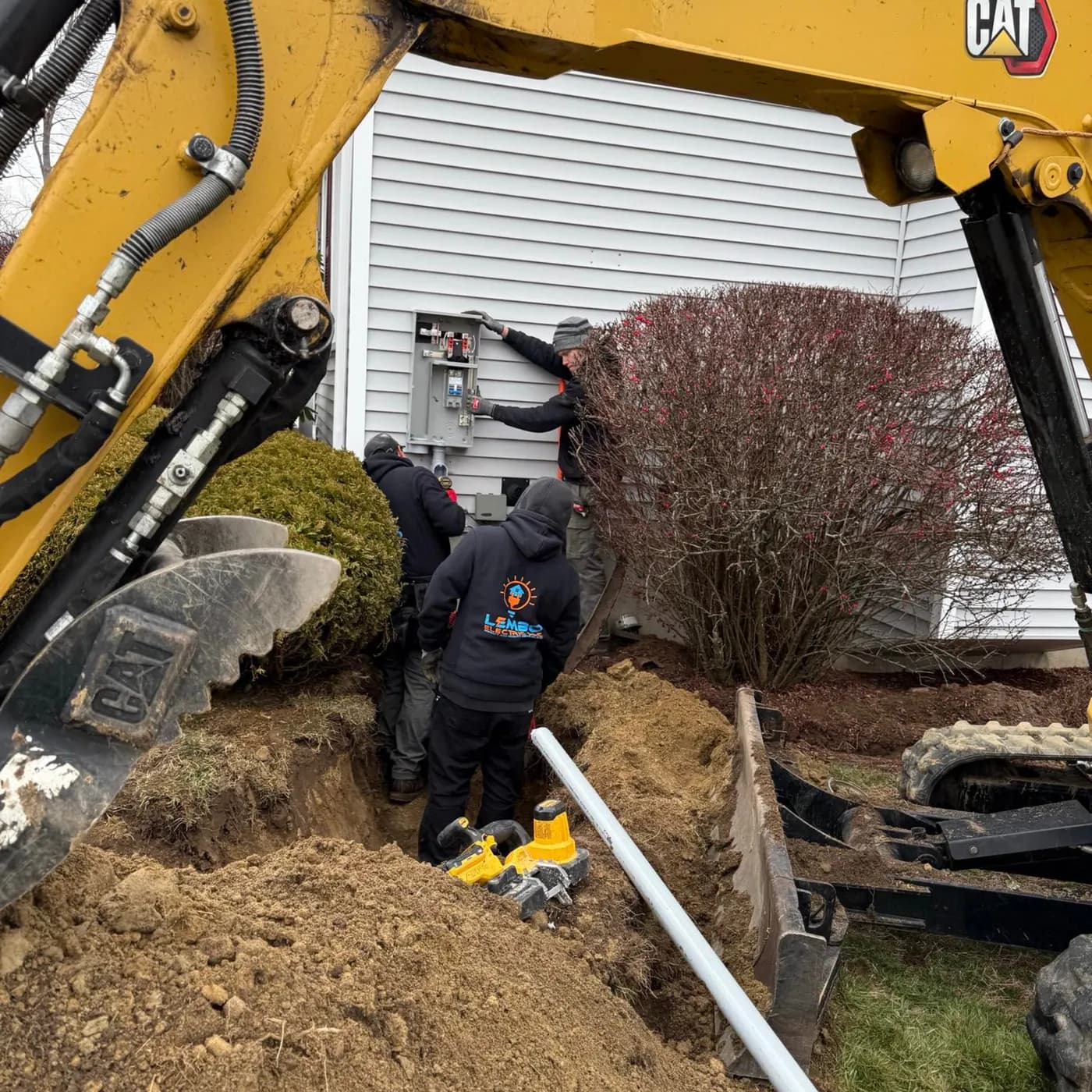 Four crew members in dark hoodies working at an exterior meter and disconnect beside a dug trench, framed by the arm and bucket of a yellow mini excavator, with gray PVC conduit and power tools against gray siding.