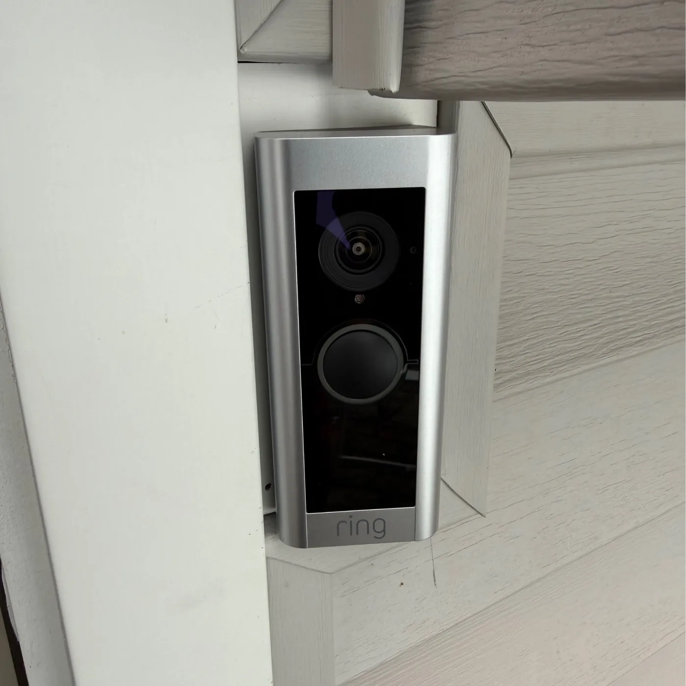 A silver Ring video doorbell on an angled mount between white trim and beige siding.