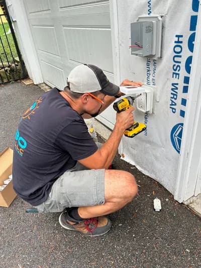 A man in a gray cap, orange safety glasses, and a dark T-shirt crouching to drill into a white electrical box mounted on house wrap beside a white garage door.