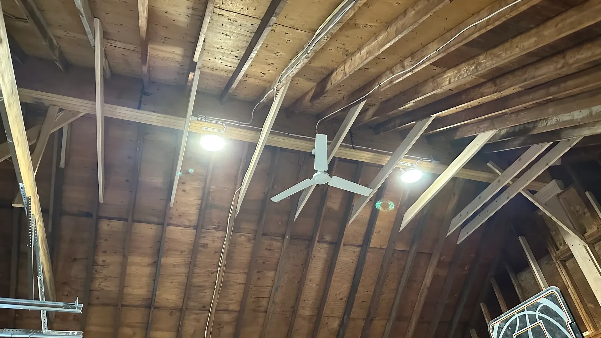 Wood barn rafters with two lit round LED fixtures, a white three-blade ceiling fan, and a basketball hoop at the lower right.