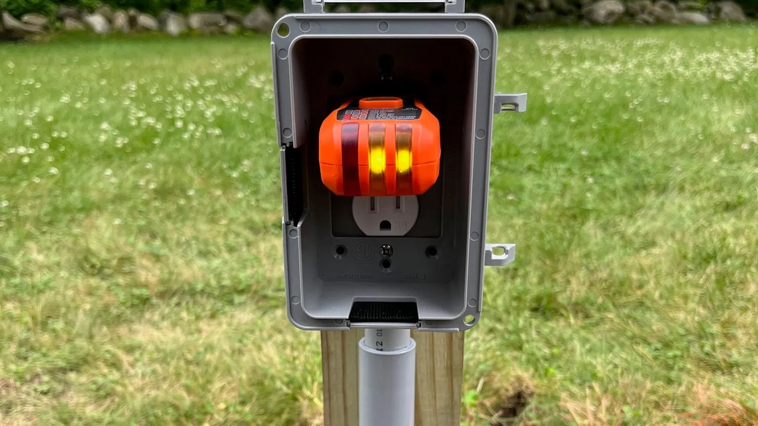 An orange plug-in outlet tester in an open gray weatherproof in-use box on a wood post, with gray PVC conduit running down the post and lawn and orange marker flags behind it.
