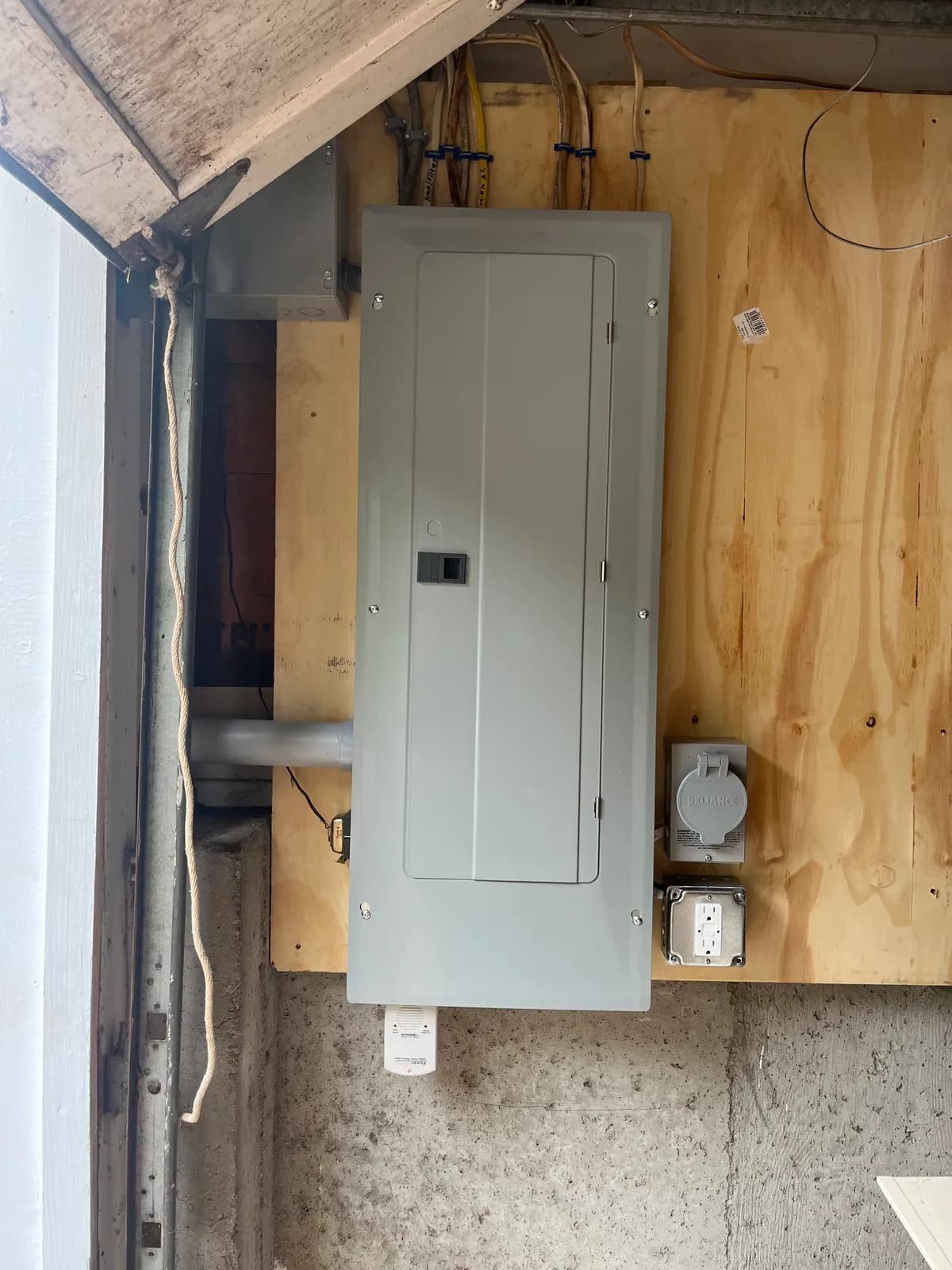 A gray breaker panel on a plywood backboard beside a garage door track, with a Reliance power inlet box and a GFCI receptacle in a metal box at its lower right and cables running into the top.