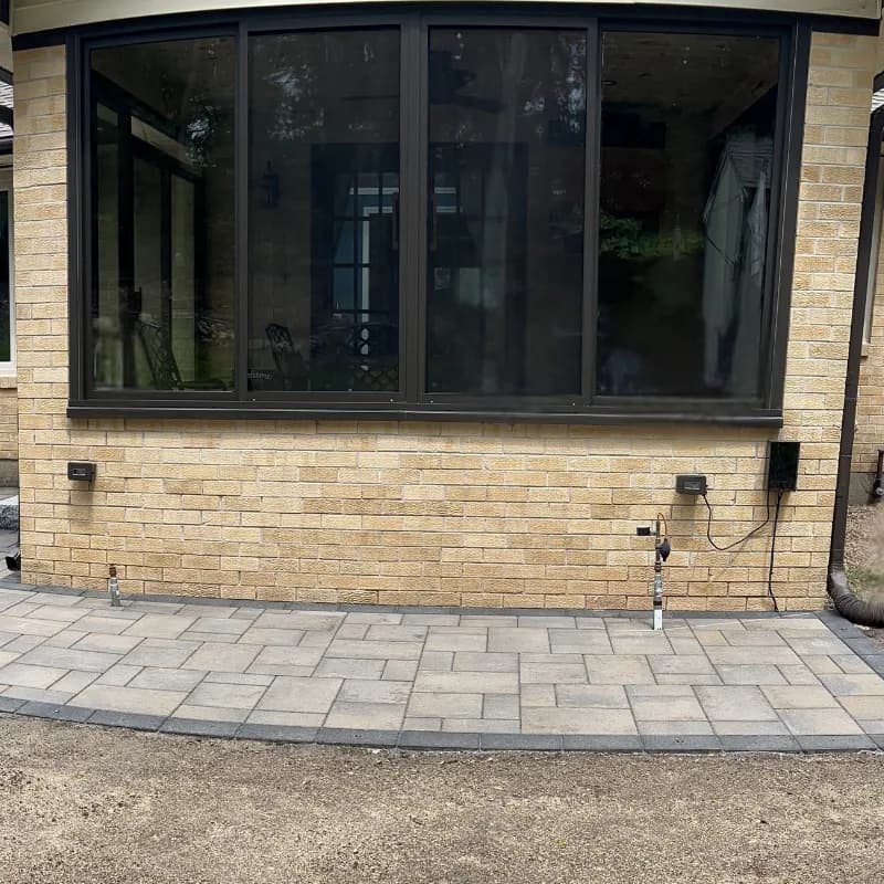 Three gray weatherproof in-use outlet covers and a hose bib on a tan brick wall below large black-framed windows, above a paver patio.