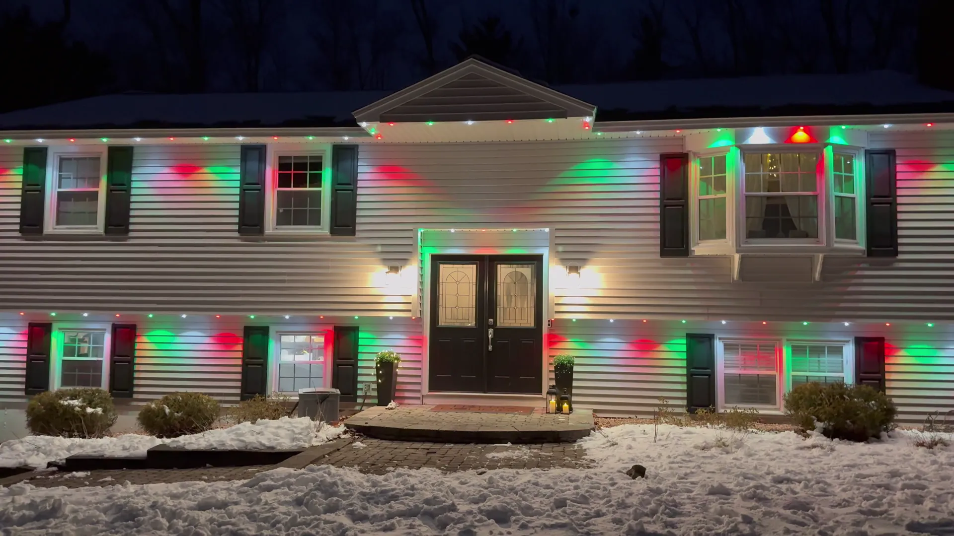 A split-level house at night after a snowfall, with red, green, and white permanent lights along the eaves washing the white siding, black shutters, and double front doors lit by two sconces.