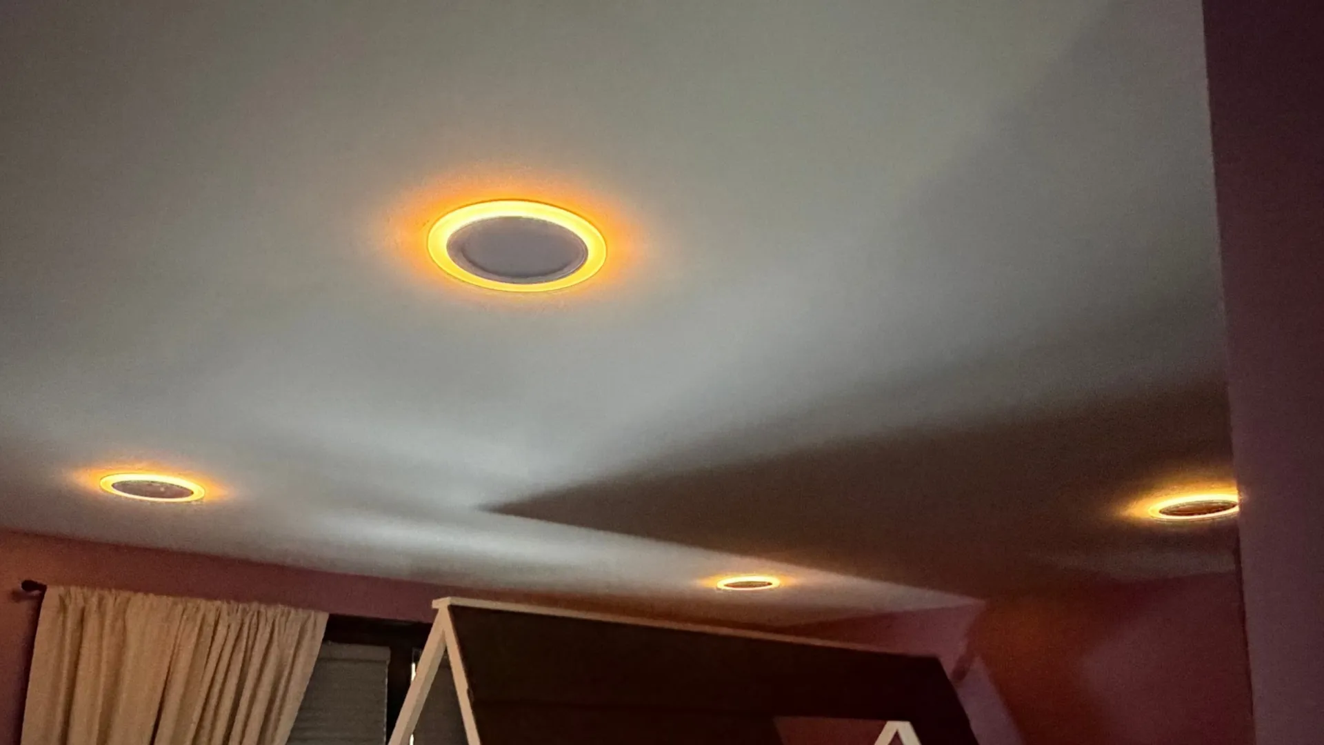 A purple kid's bedroom with a playhouse loft bed under a dark roof, recessed ceiling lights glowing with warm orange halo rings, and a curtained window.