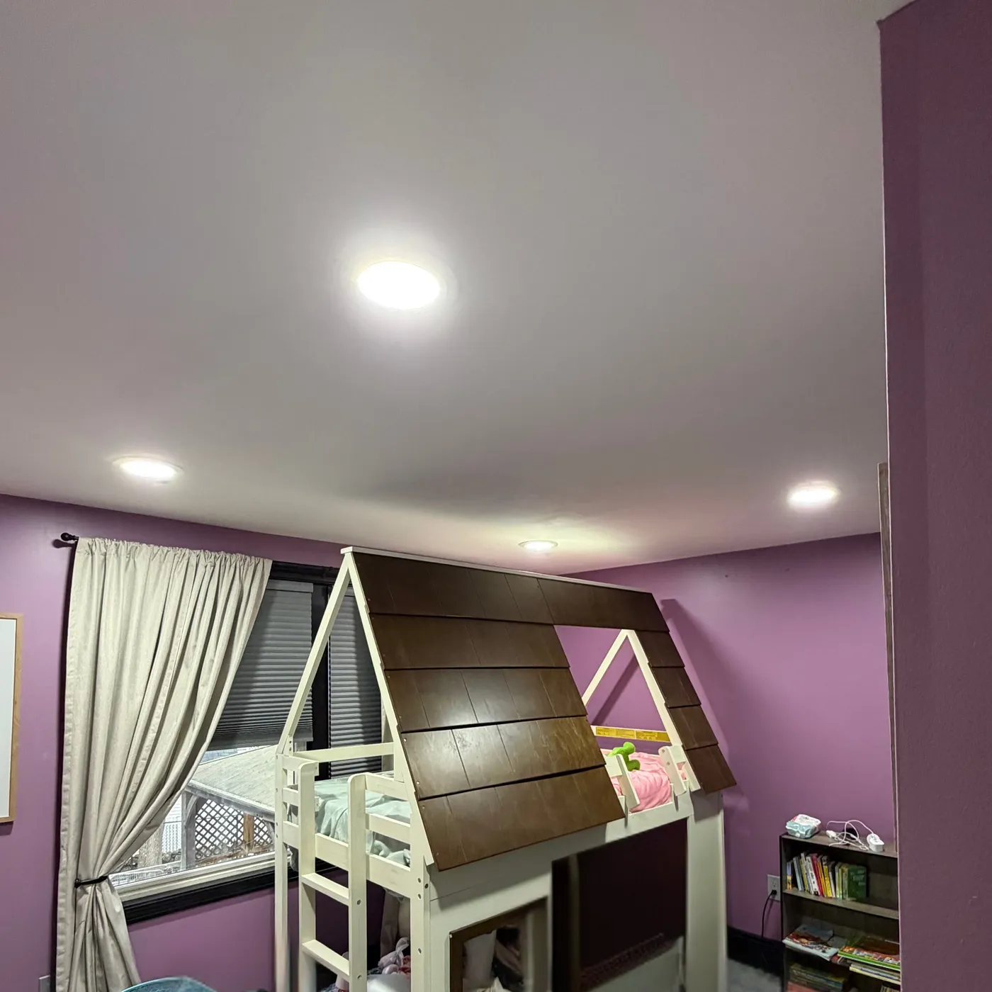 A purple kid's bedroom with a playhouse loft bed and a bookshelf, lit bright white by recessed ceiling lights.