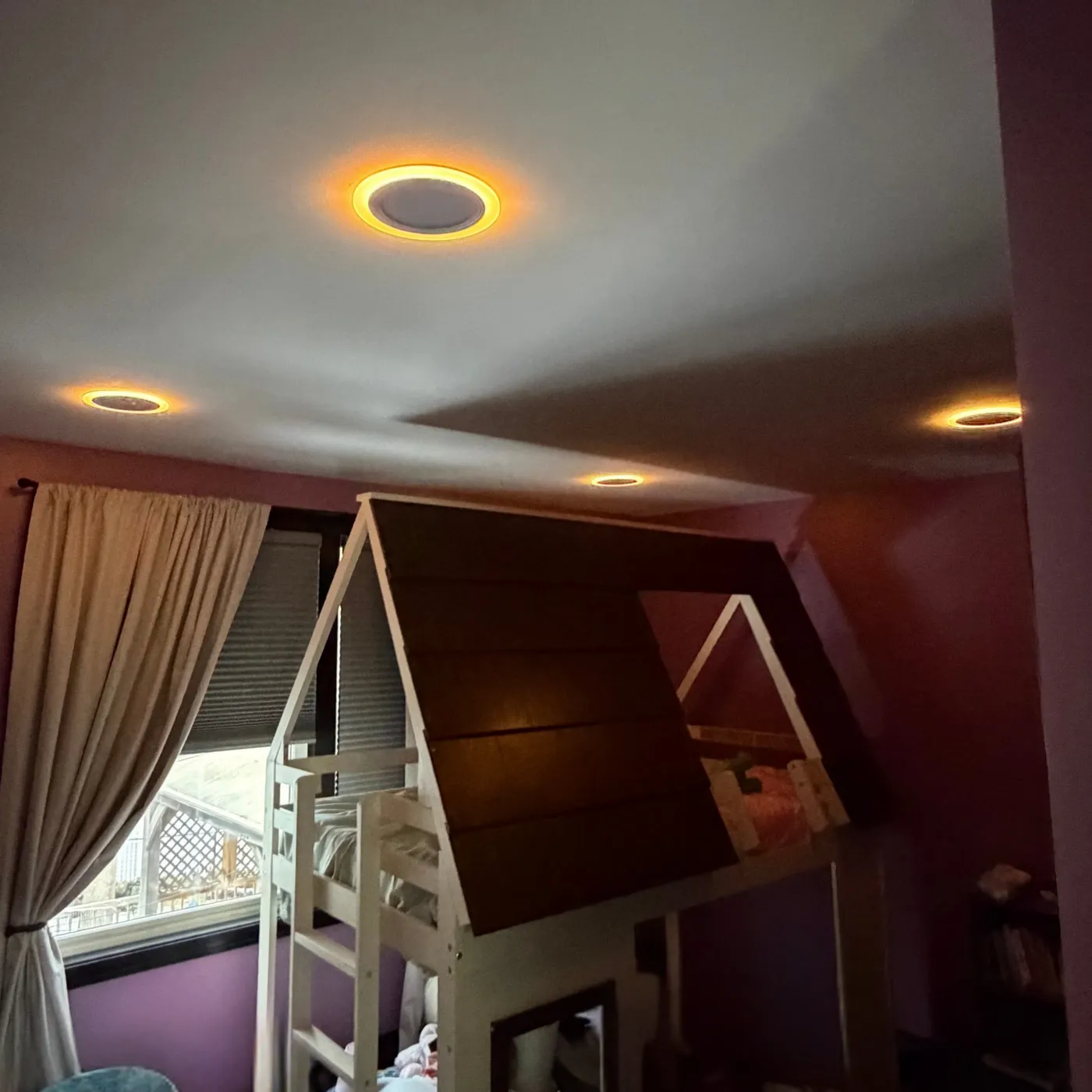 A purple kid's bedroom at night, lit only by the warm orange night light rings of recessed ceiling lights above a playhouse loft bed and a curtained window.