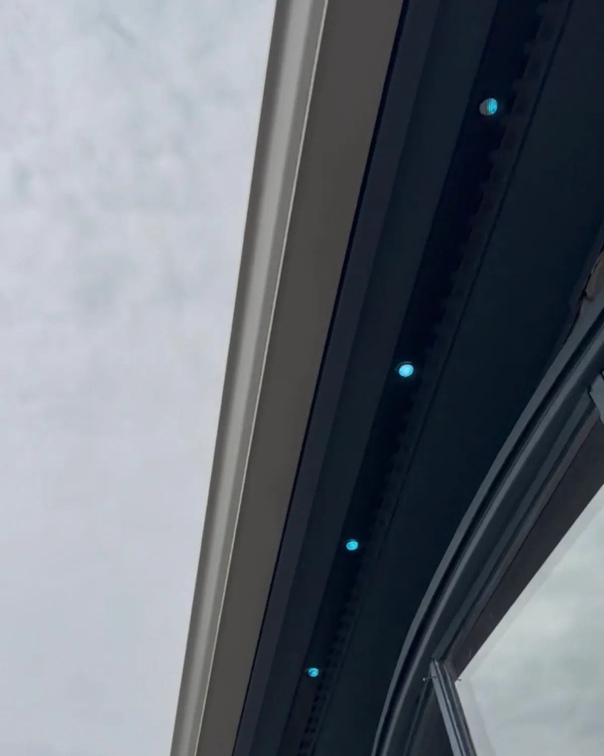 Looking up at a white gutter and dark soffit where a mounting track holds a row of round LED lights glowing teal, with a curved dark-framed window at the right under an overcast sky.