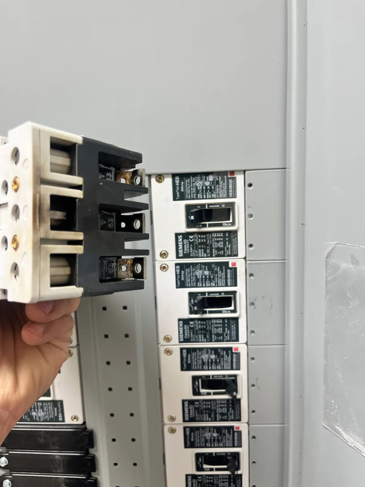 A hand holding a large three-pole circuit breaker beside an open panel lined with rows of Siemens breakers.
