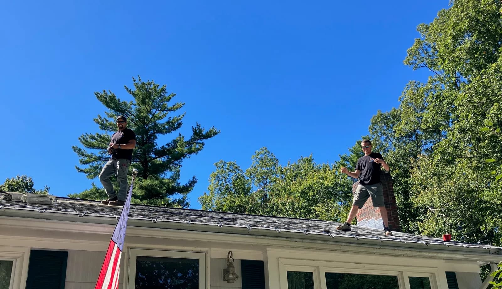 Two men in black T-shirts standing on the gray shingled roof of a ranch house under a blue sky, with a tall pine behind them and an American flag on the porch below.