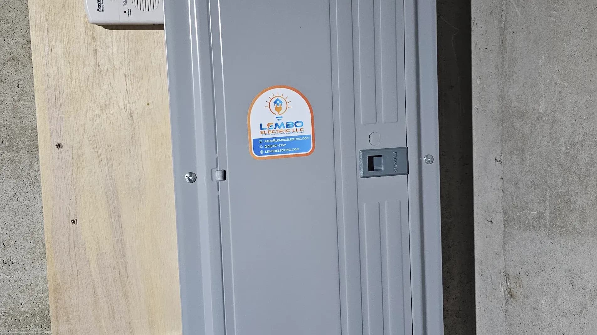 A gray electrical panel mounted on a plywood backer in a basement, with a round Lembo Electric sticker on its door.