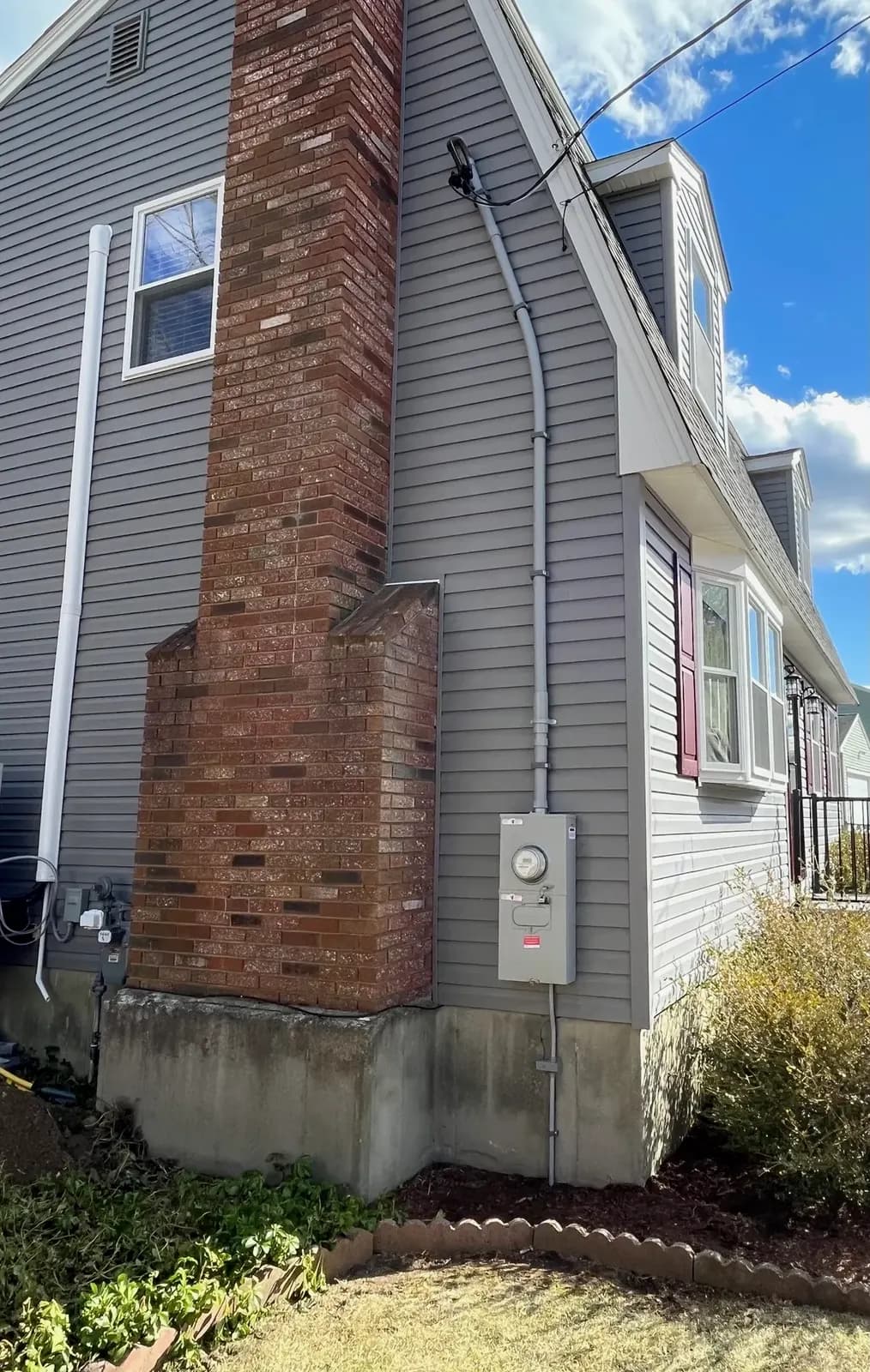 Side of a gray-sided cape with a brick chimney, a new meter socket with main disconnect and a conduit mast running up the wall to the overhead service drop.