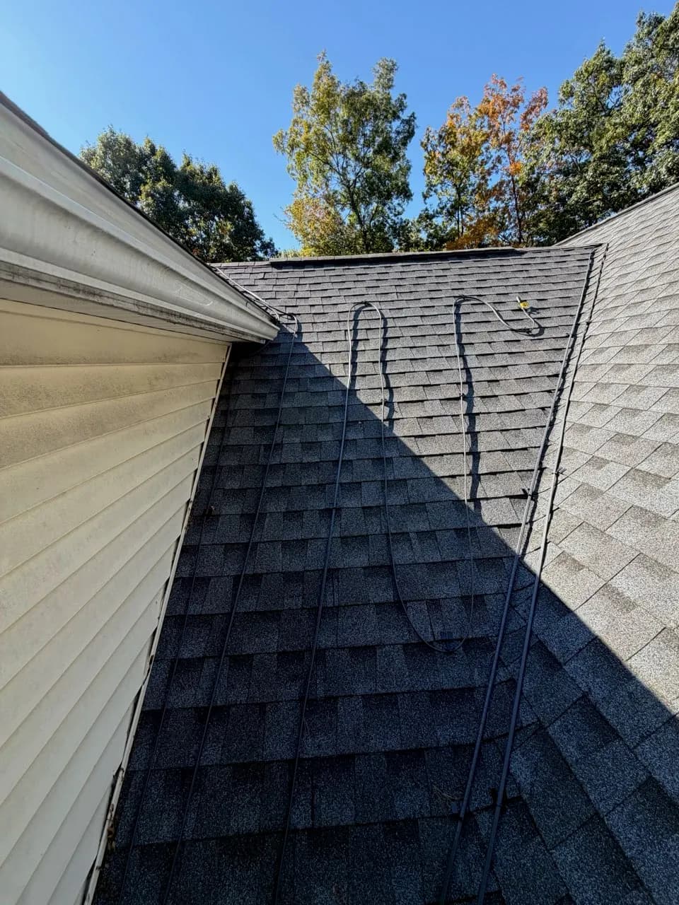 Close-up of a shingled roof slope on a sunny day with black heat cables running in long S-curves down toward the eave, beside a cream vinyl-sided wall.