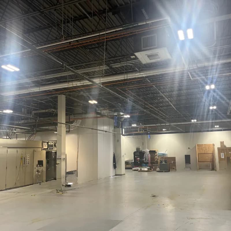 A large open commercial space with a pale concrete floor and white walls, lit by LED fixtures hung from a dark exposed-steel ceiling with ductwork and conduit.