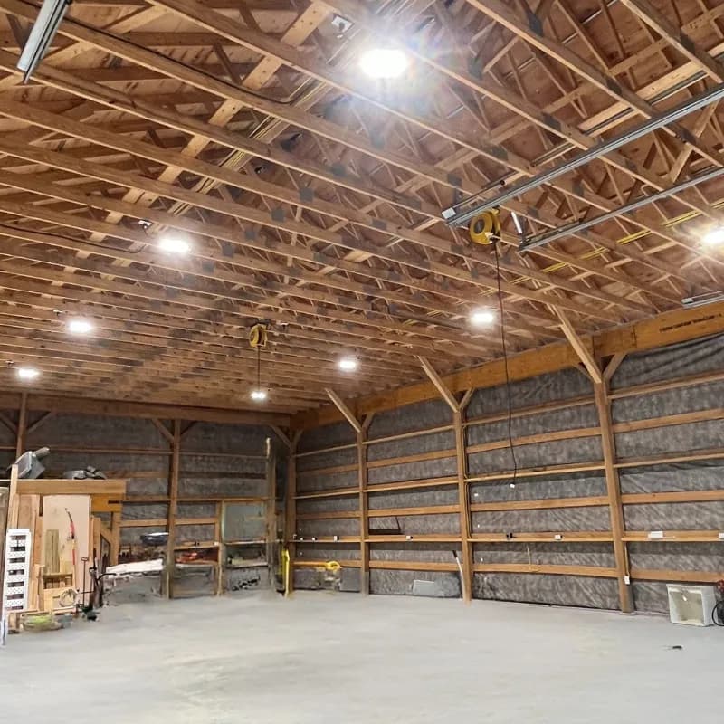 The inside of a large timber-framed building with round LED fixtures glowing across an open wood-truss ceiling above an empty concrete floor.