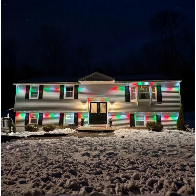 A raised ranch home at night after a snowfall, its eaves lined with permanent LED lights washing the white siding in alternating red and green.