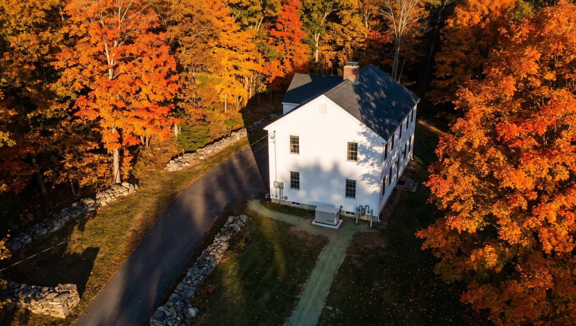 Aerial view of a white farmhouse at the end of a paved driveway, ringed by stone walls and orange and red autumn maples.