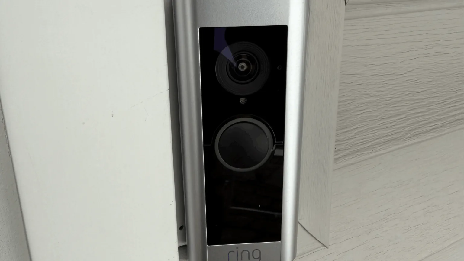 A silver Ring video doorbell mounted flush on the white trim beside a home's front entry.