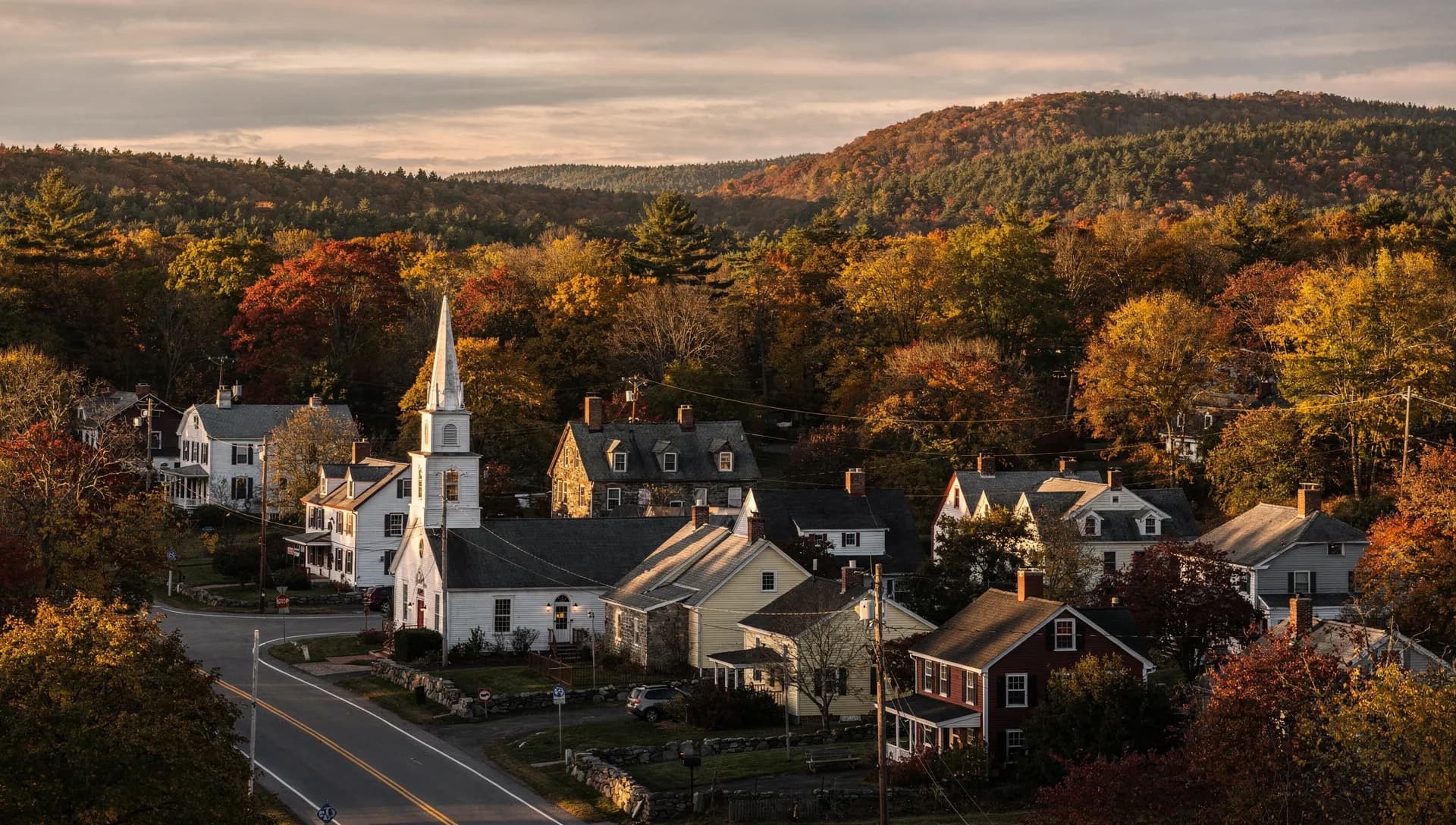 An elevated golden-hour view over a New England village with a white church steeple, Colonial and Cape homes and autumn hills rolling to the horizon.
