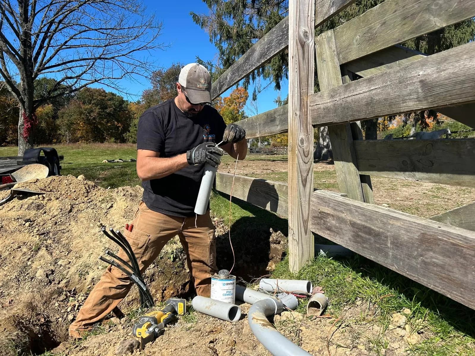 A Lembo Electric technician in a company cap and work gloves gluing PVC conduit at a fence-line trench on a clear fall day.