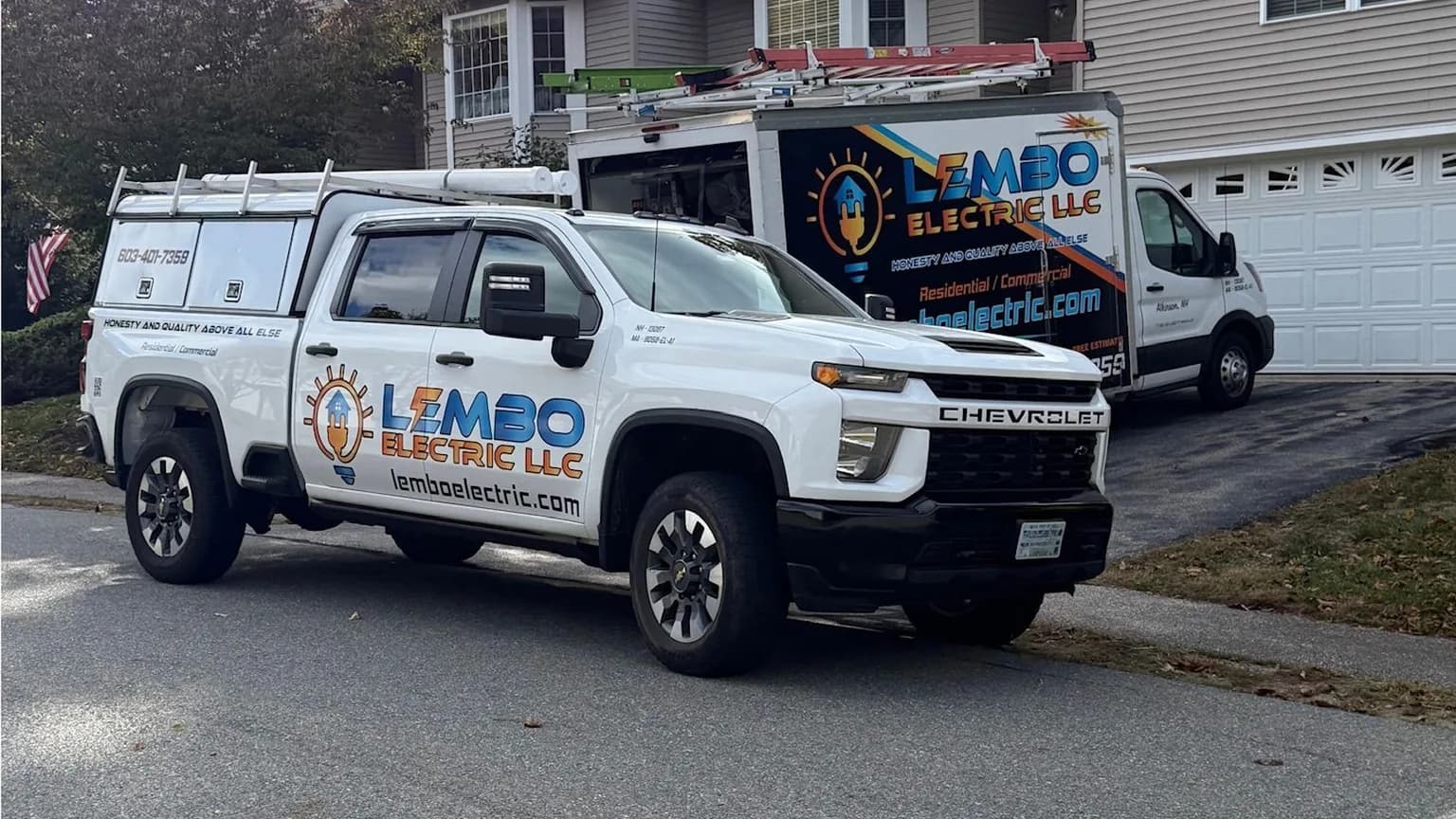 A white Lembo Electric pickup with a ladder rack parked at the curb and the lettered Lembo Electric box van in the driveway of a gray house on a sunny fall day.