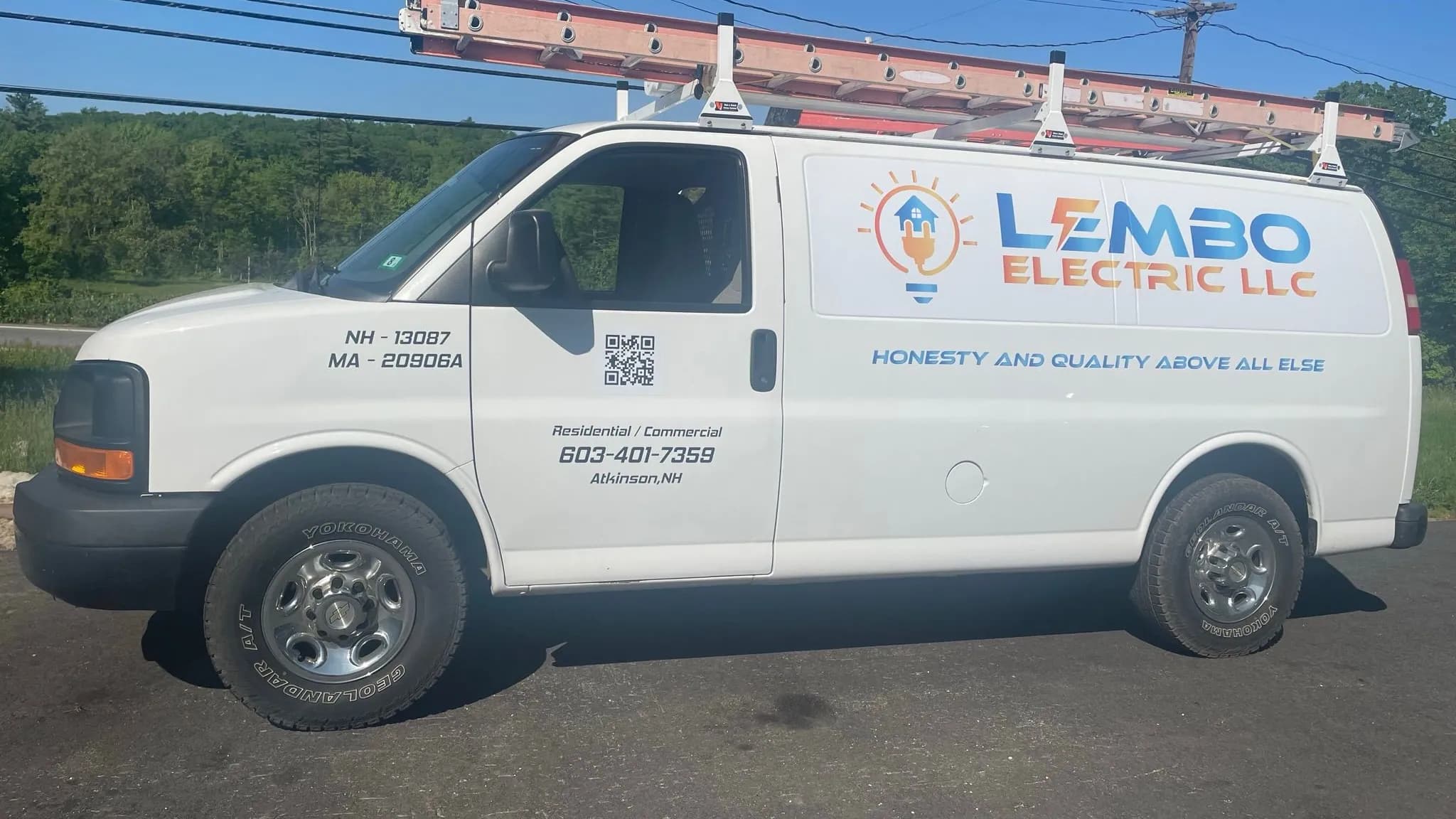 A white Lembo Electric LLC work van with an orange extension ladder on its roof rack, parked on a paved lot beside green summer trees under a clear blue sky.