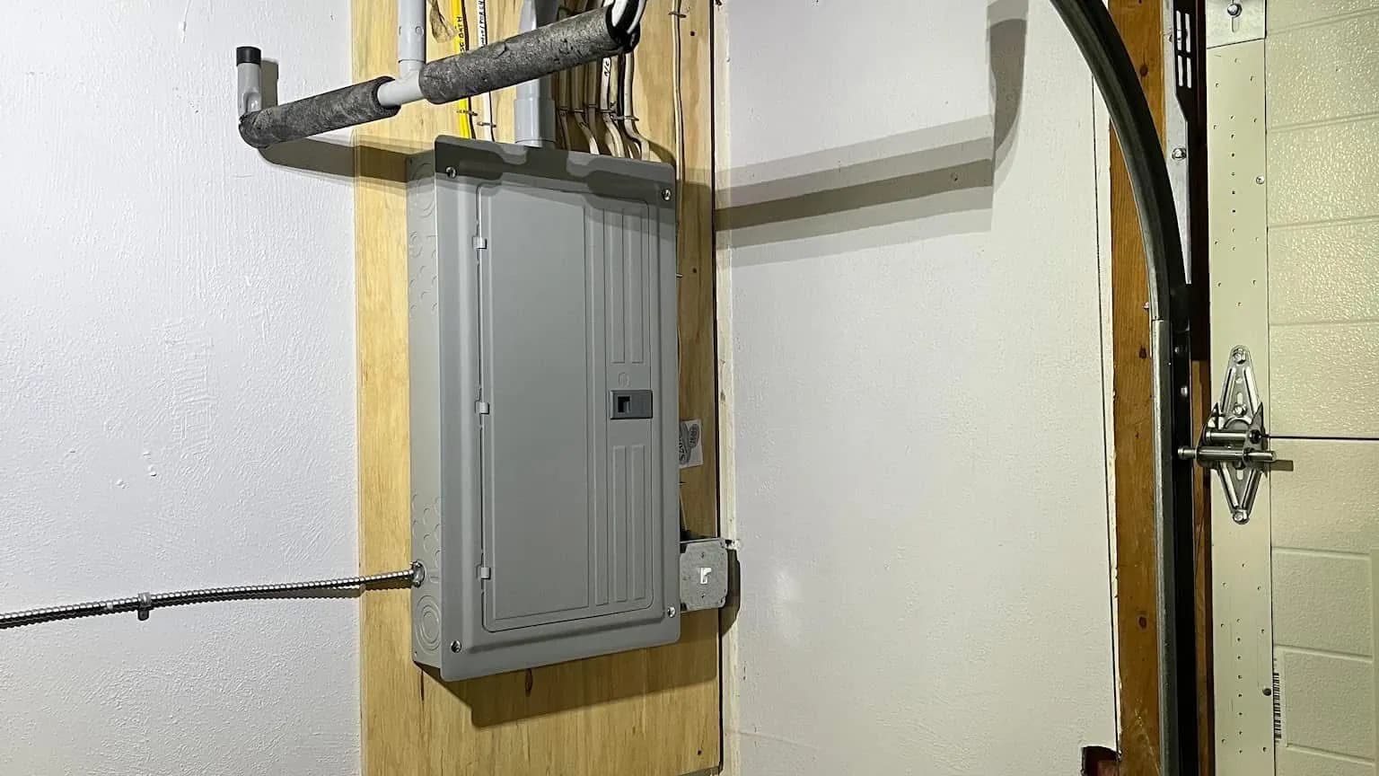 A gray breaker panel on a plywood backboard in a garage beside the overhead door track, with PVC conduit and insulated pipe above and flexible metal conduit feeding the side.