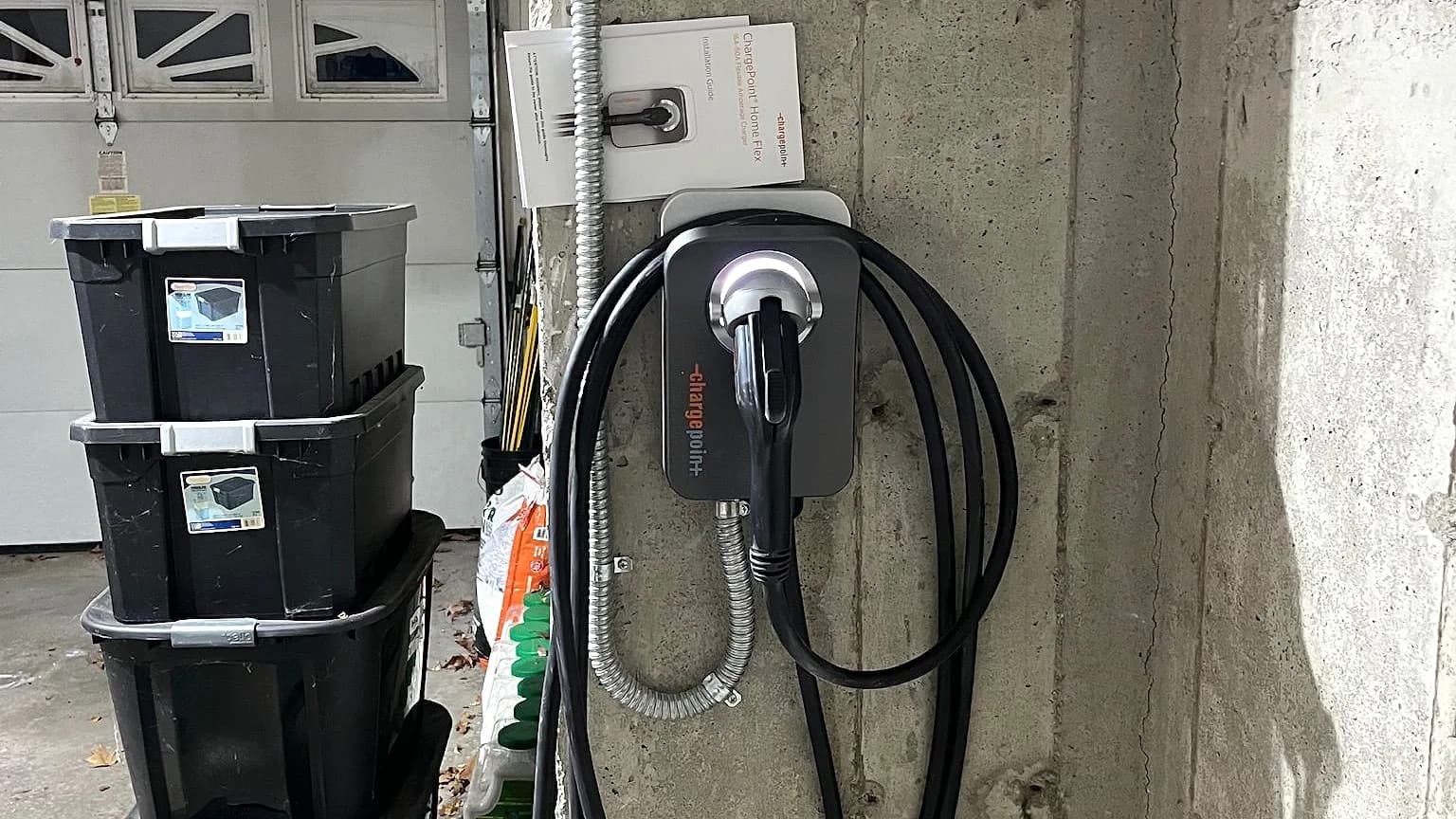 A ChargePoint Home Flex charger hardwired through flexible metal conduit on a concrete garage pillar, its black cable coiled around the unit and the installation guide tucked above it, storage bins and the garage door beside it.