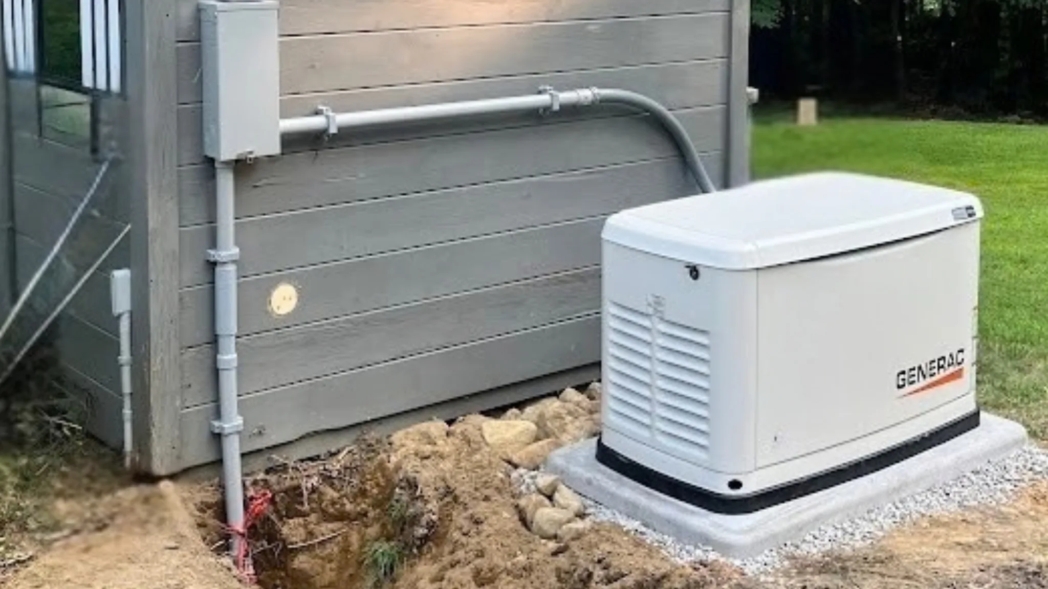 A Generac standby generator on a new concrete pad beside a gray-sided shed, with conduit to a disconnect box on the shed wall and an open trench in front in late-summer light.