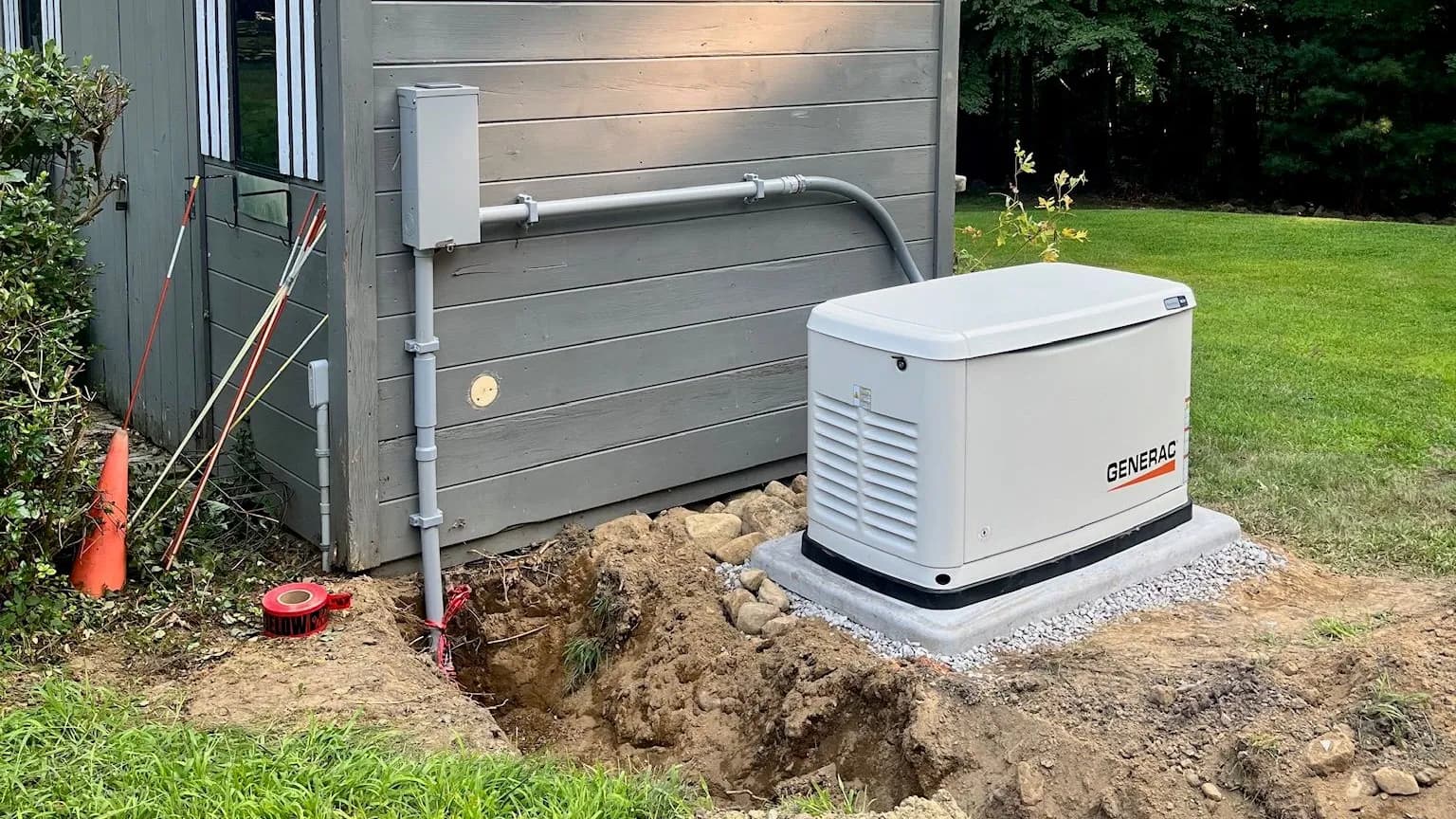A Generac standby generator on a new concrete pad beside a gray-sided outbuilding, with conduit to a disconnect box, an open trench and a roll of red warning tape in late-summer light.
