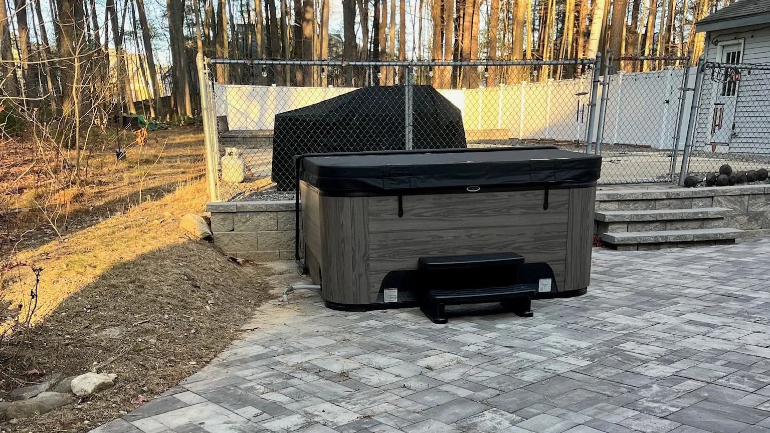 A covered hot tub with steps on a gray paver patio, beside a chain-link fence and a covered grill, in low golden light among bare trees.