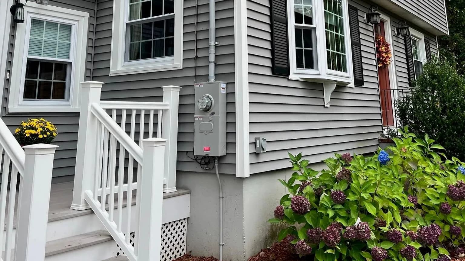 A gray meter socket with meter and conduit on gray vinyl siding beside white porch stairs, with blue and purple hydrangeas, potted mums and an autumn wreath on the red front door.