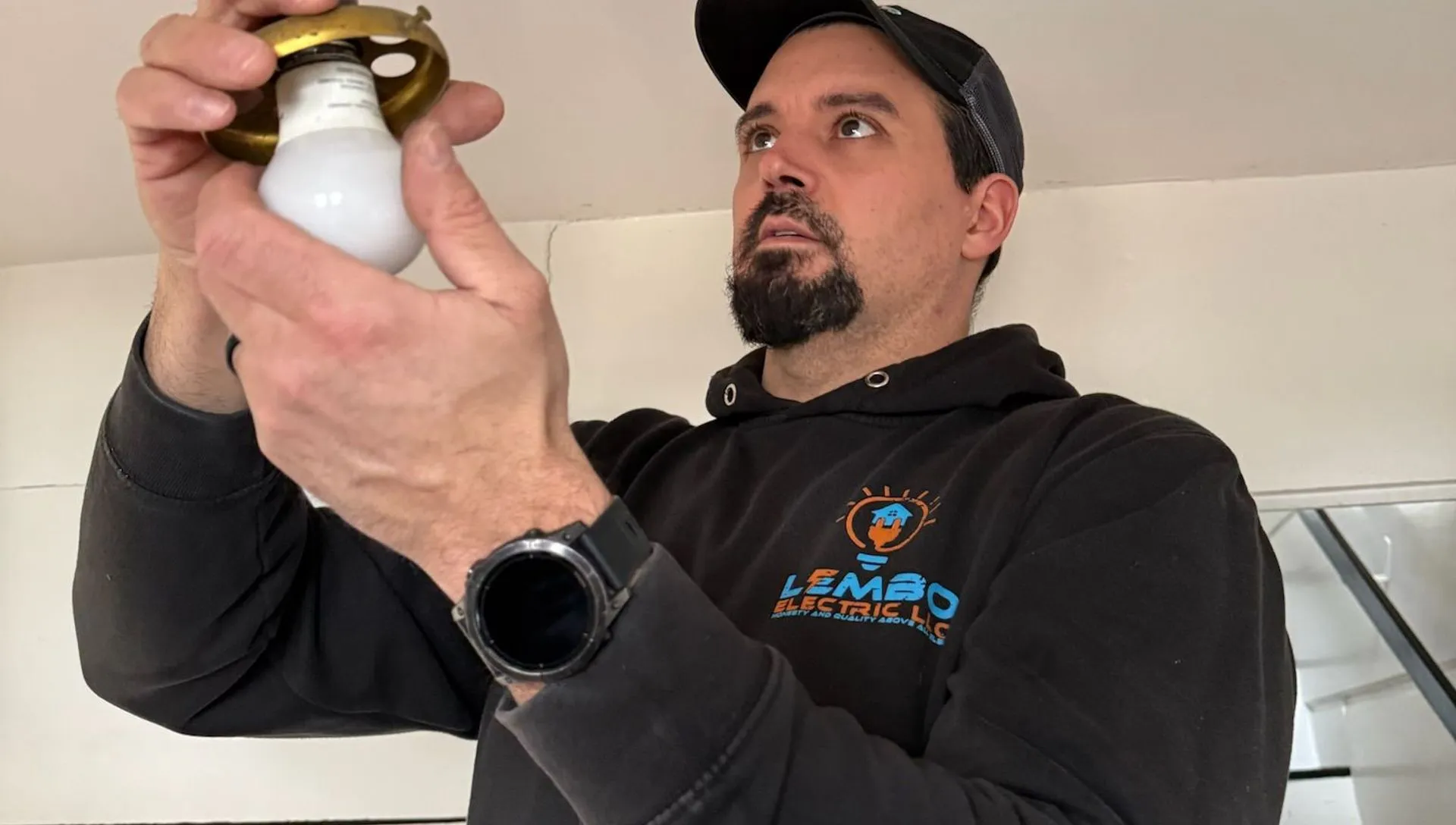 A Lembo electrician in a branded hoodie and cap inspects a vintage brass chain-hung light fixture with a bulb in an older home.