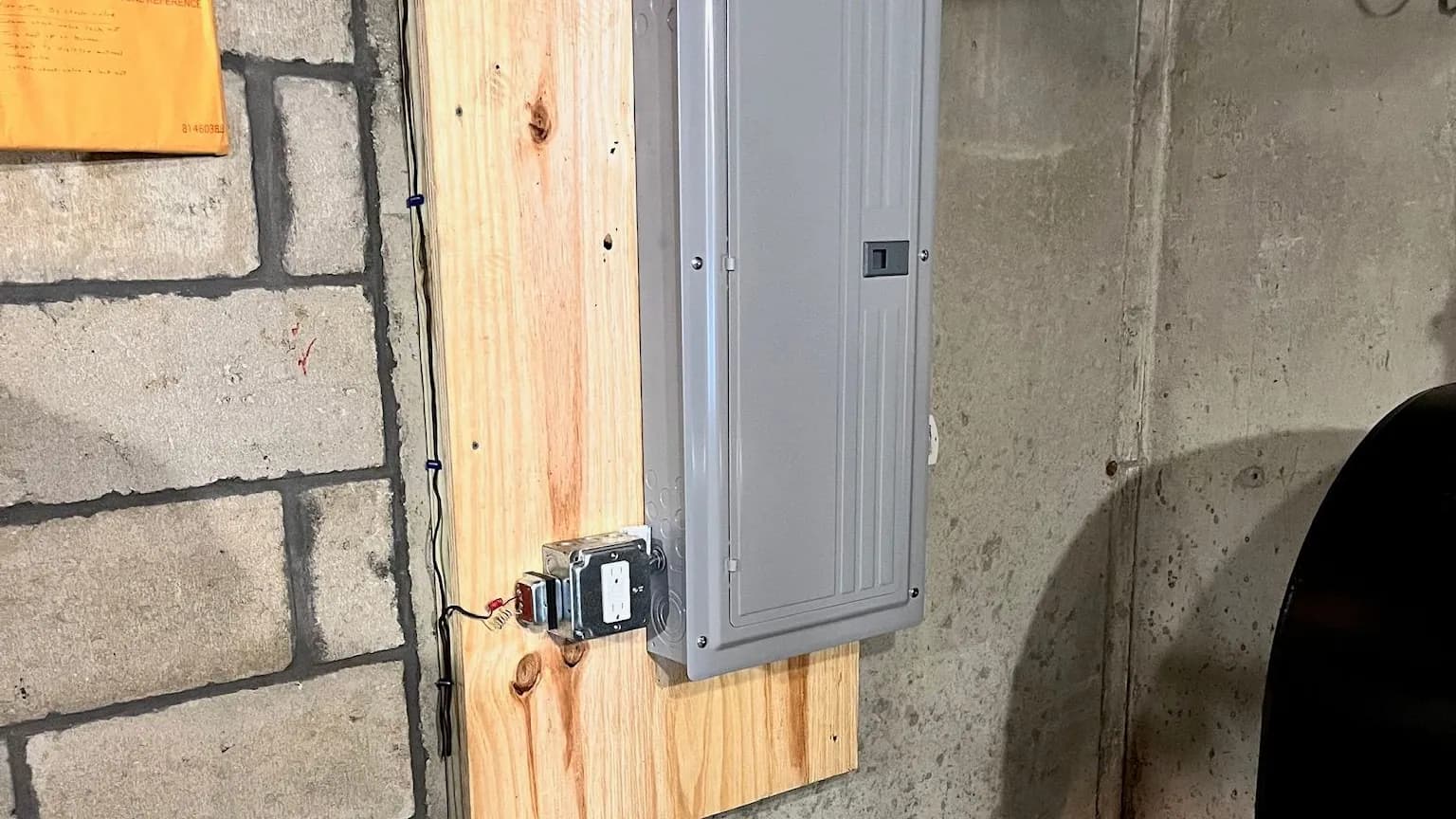 A new gray breaker panel with its door closed, mounted on a pine backboard in an unfinished basement, with a GFCI outlet in a metal box beside it.