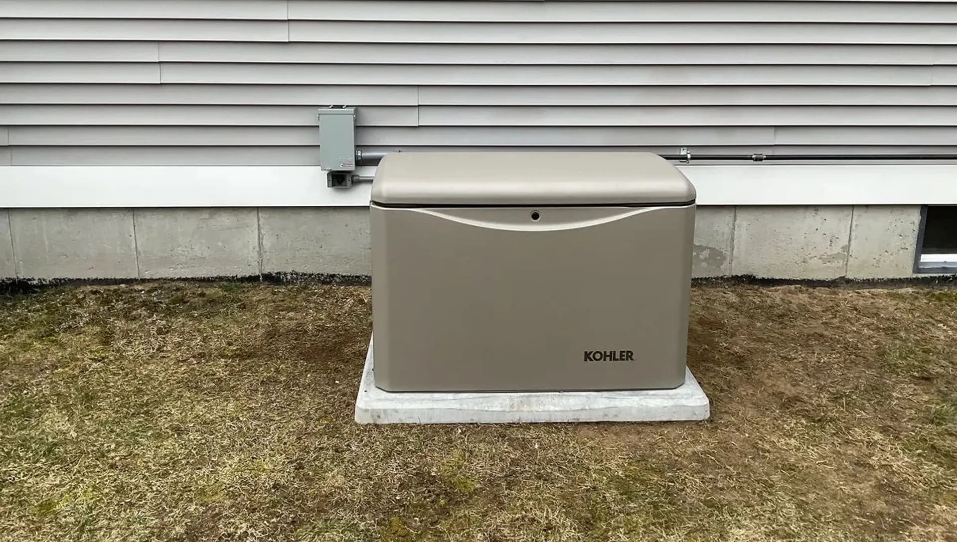 A Kohler home standby generator on a concrete pad against light gray siding, with a disconnect box and conduit running along the foundation line.