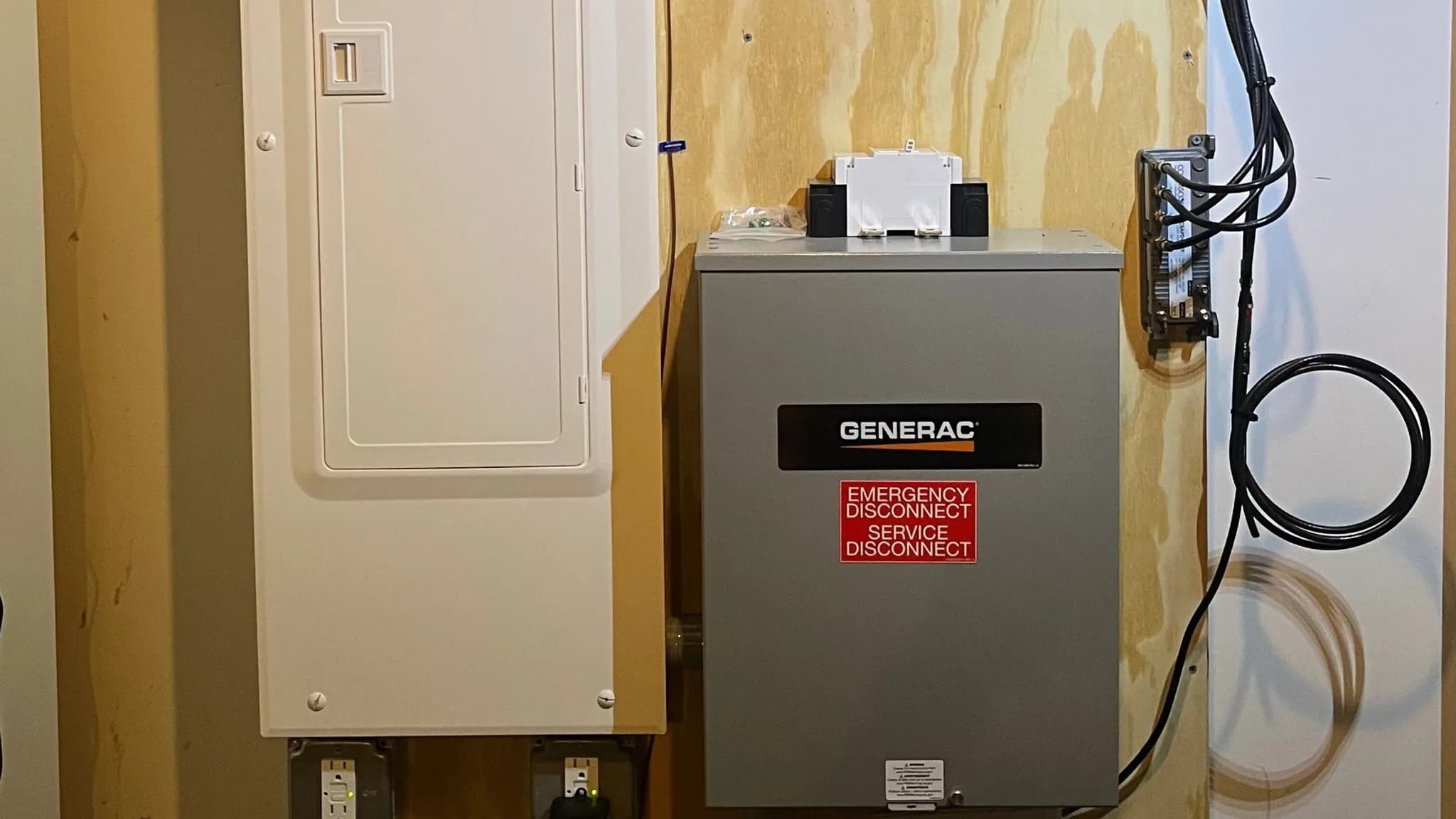 A white breaker panel beside a gray Generac automatic transfer switch marked emergency service disconnect, both mounted on a plywood backboard with cables coiled at the right.