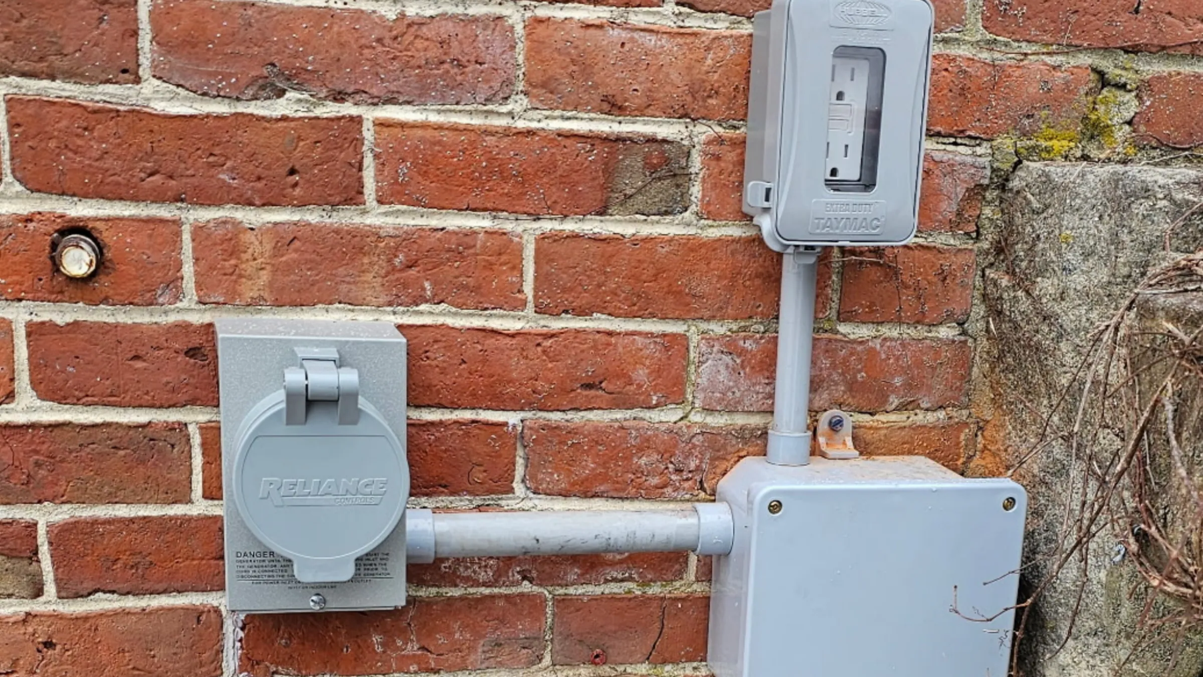 A Reliance Controls generator power inlet box and a Taymac covered GFCI outlet, linked by gray PVC conduit to a junction box on an old red brick wall.
