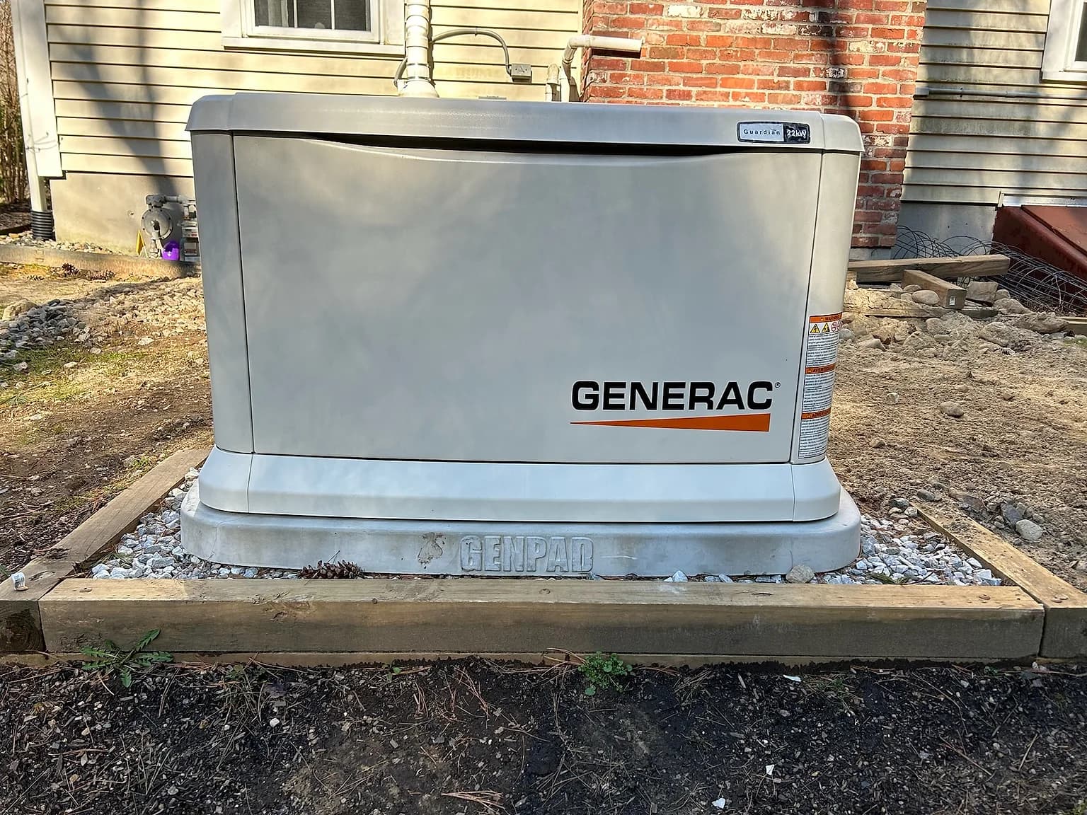 A new Generac Guardian 22kW standby generator on a Genpad base inside a timber-framed bed of white stone, beside a brick chimney and tan vinyl siding.