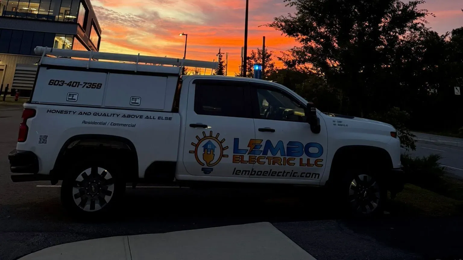 A white Lembo Electric pickup with a ladder rack parked by an office building under an orange and pink sunset, a street light glowing against the sky.