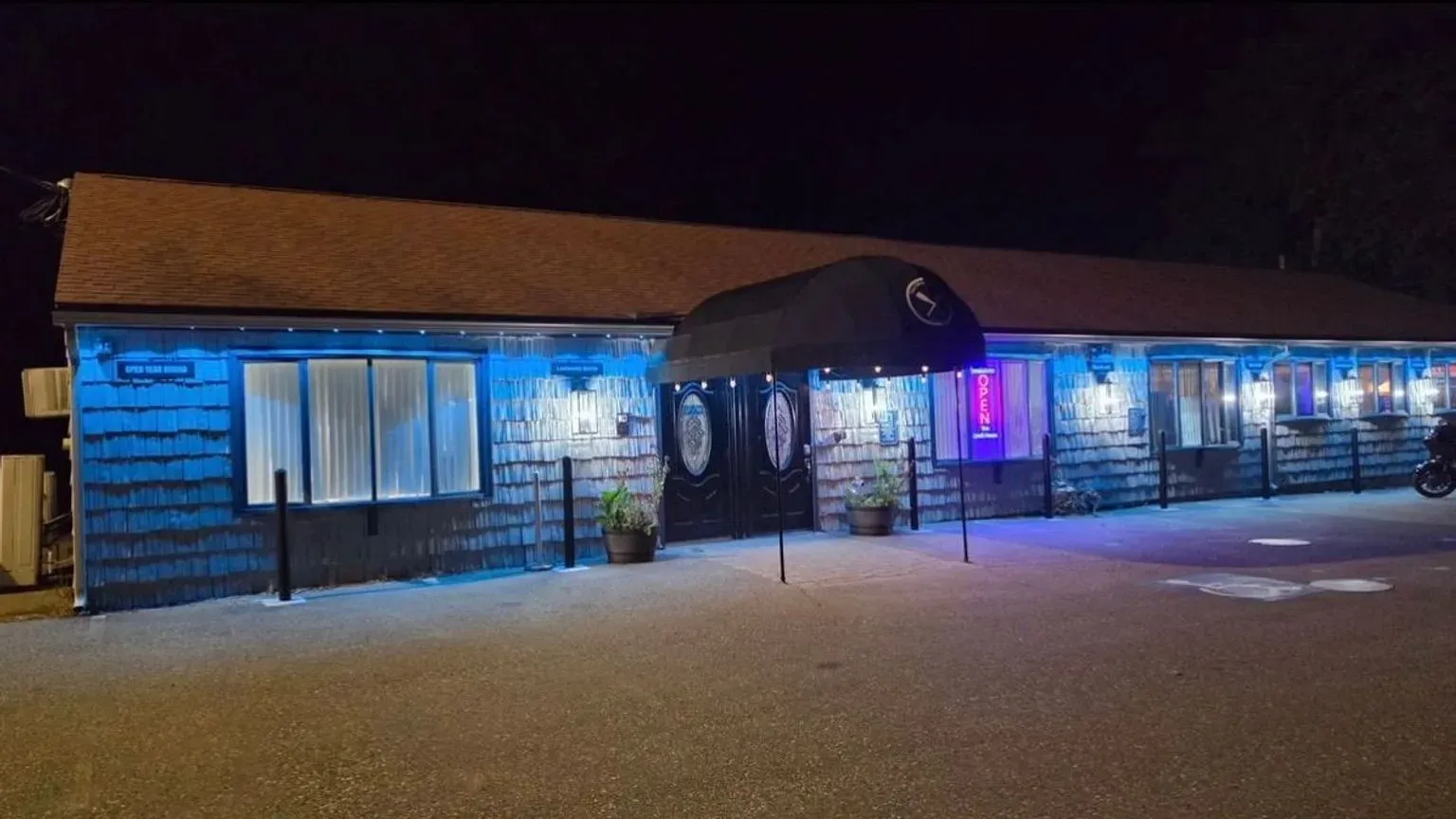 A long single-story commercial building at night, its shingled front washed in blue by permanent eave lights, with an entry awning, lit wall sconces and a glowing open sign.