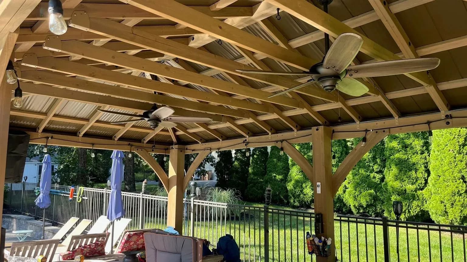 A cedar backyard pavilion with two outdoor ceiling fans and hanging bulb lights along the beams, patio seating below and a fenced pool yard with loungers and umbrellas beyond.