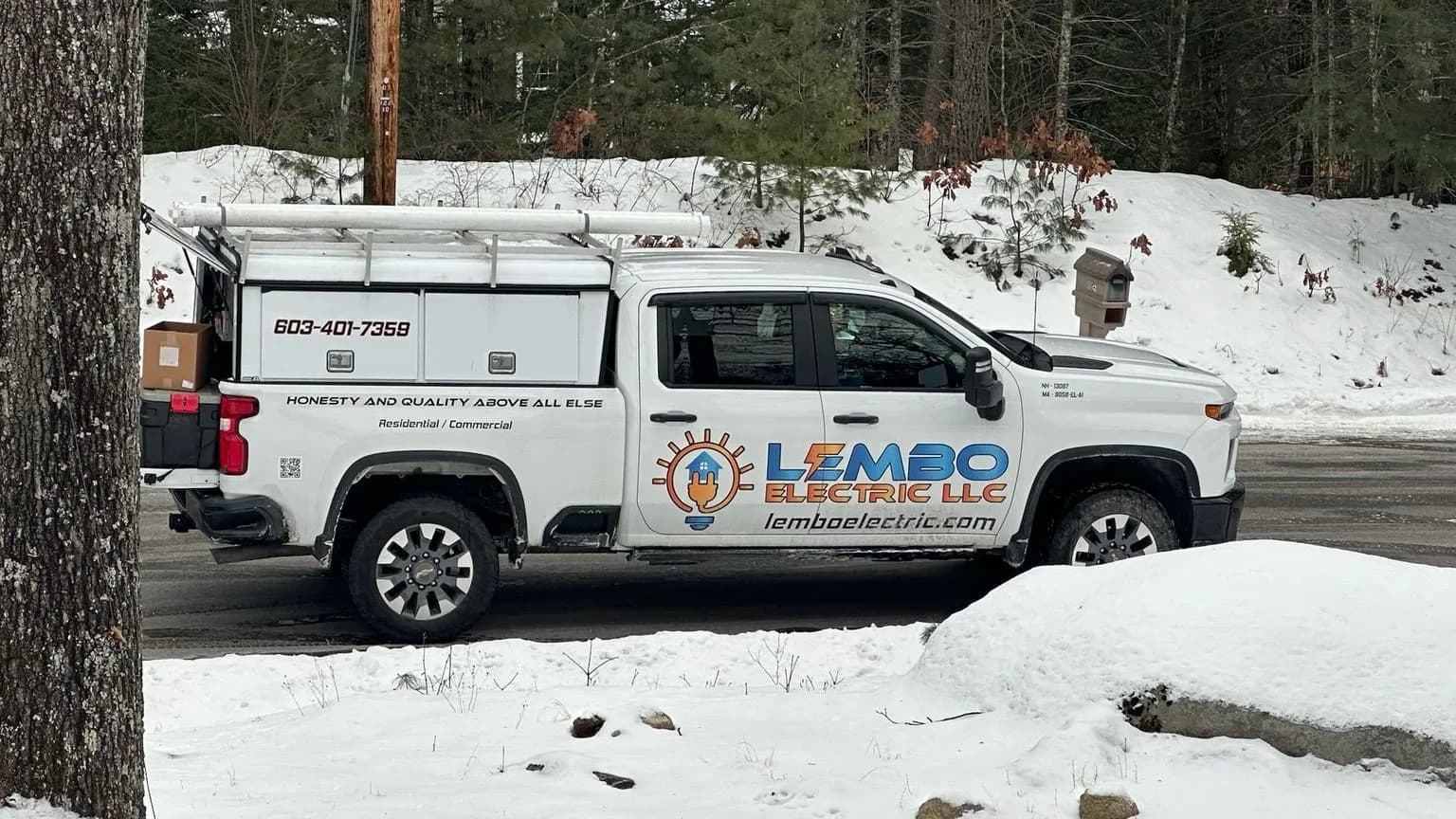 A white Lembo Electric LLC pickup with a utility cap and ladder rack, lettered with the slogan, phone number and website, parked on a snowy wooded street.