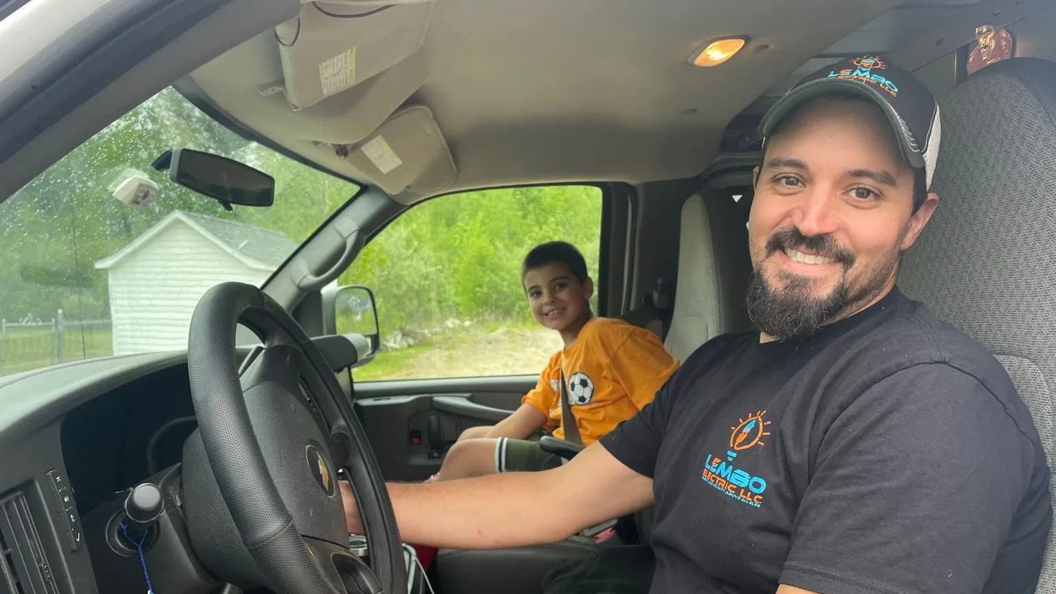 A smiling Lembo electrician in a logo shirt and cap sits behind the wheel of a work van while a boy in an orange soccer jersey rides in the passenger seat.