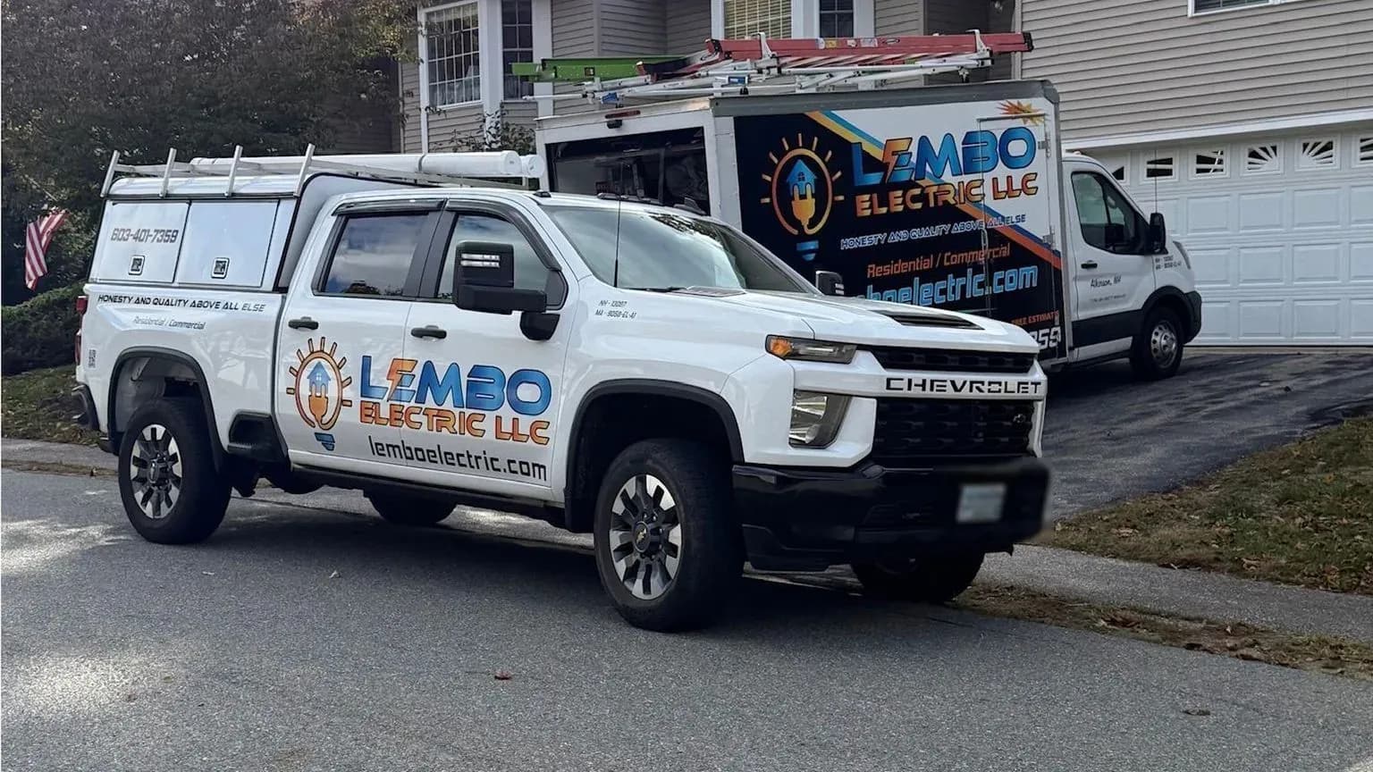 A white Lembo Electric pickup with a ladder rack at the curb and the lettered Lembo Electric box van in the driveway behind it, in front of a gray house on a fall day.