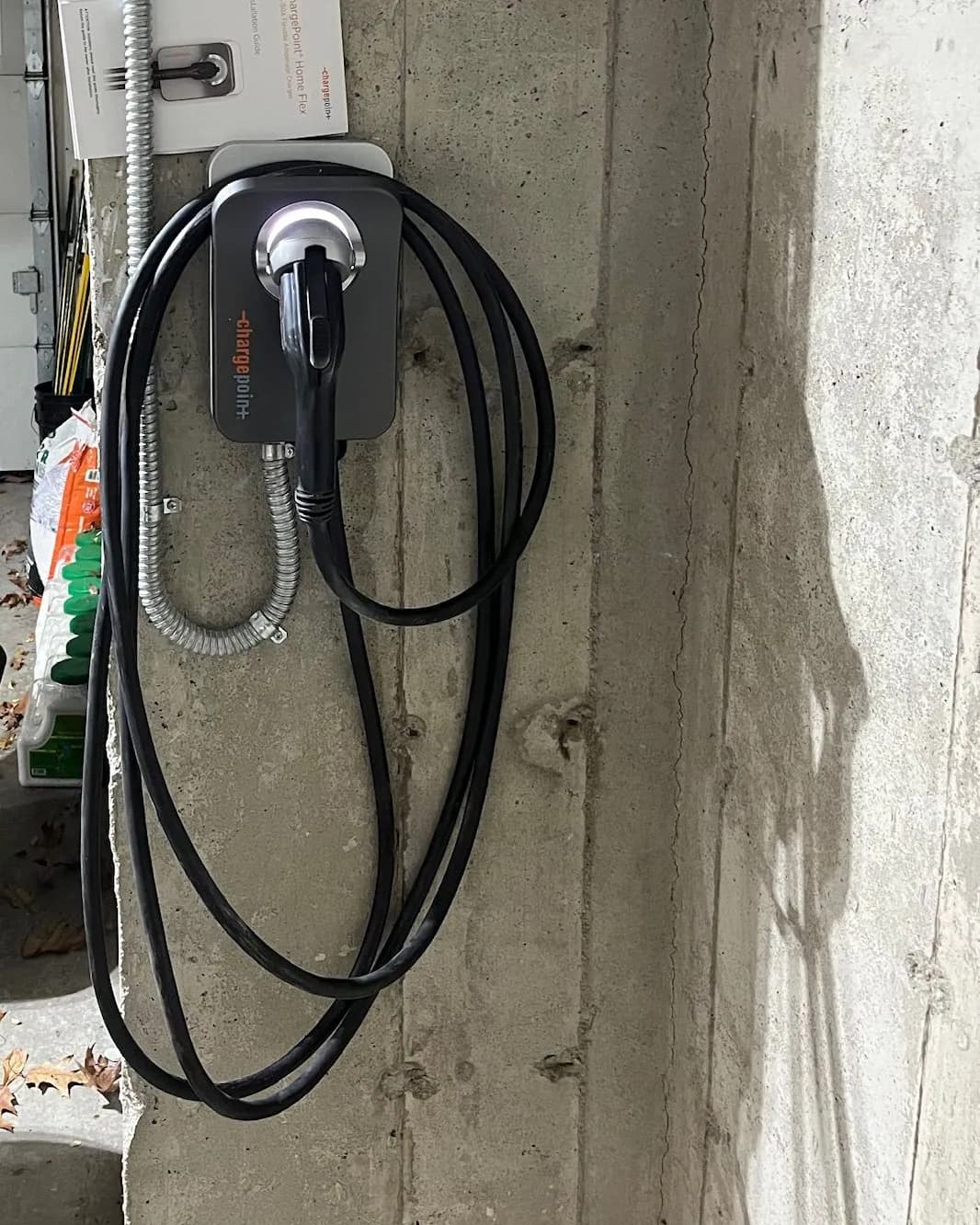 A ChargePoint Home Flex EV charger with its coiled cable, mounted on a concrete garage pillar and fed by flexible metal conduit, with fallen leaves on the floor.