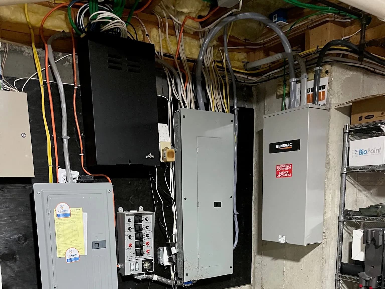 A basement electrical wall with a Lembo Electric-labeled panel, a manual transfer switch, a main panel, a Leviton wiring enclosure and a Generac automatic transfer switch marked emergency service disconnect.