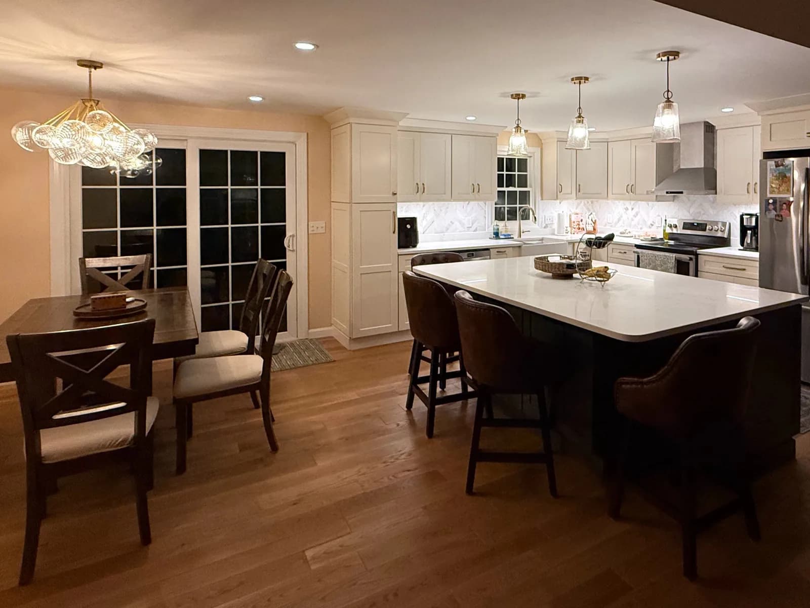 A remodeled kitchen at night with three glass pendants over a large white island with leather stools, recessed lights, under-cabinet lighting and a bubble-glass chandelier over the dining table.