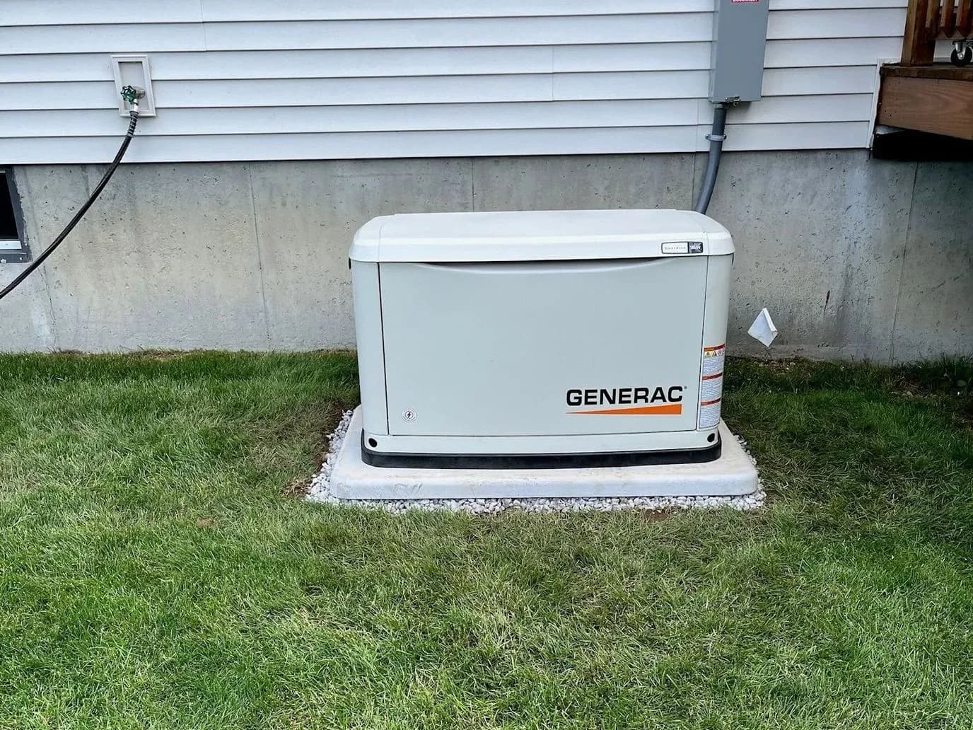 A Generac standby generator on a white pad with a gravel border, set on a green lawn against white vinyl siding and a concrete foundation, a gray disconnect box on the wall above it.