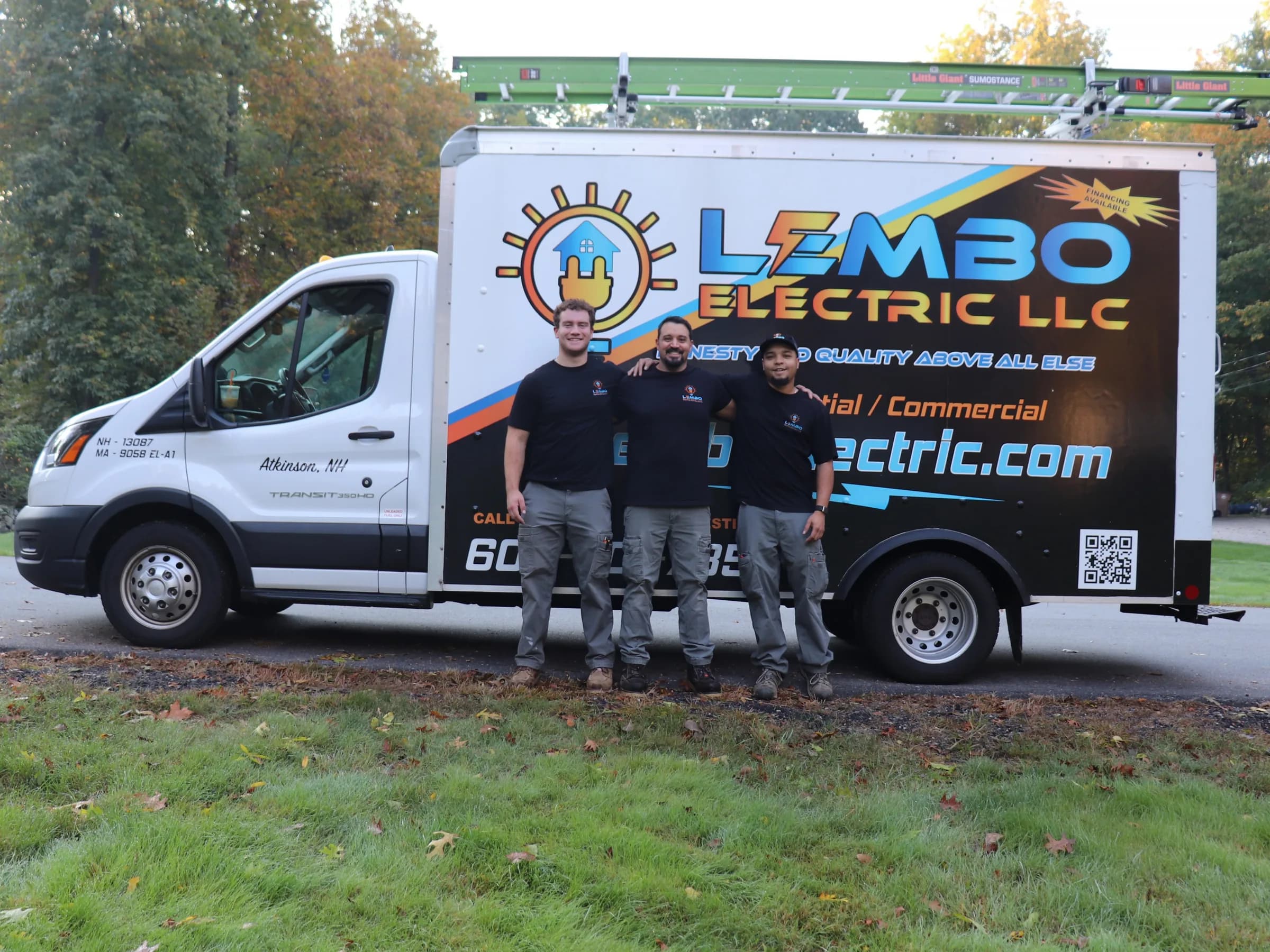 Three Lembo Electric electricians in matching black company T-shirts and gray work pants stand arm in arm in front of the lettered Lembo Electric box truck, a ladder on its roof rack and fall trees behind.